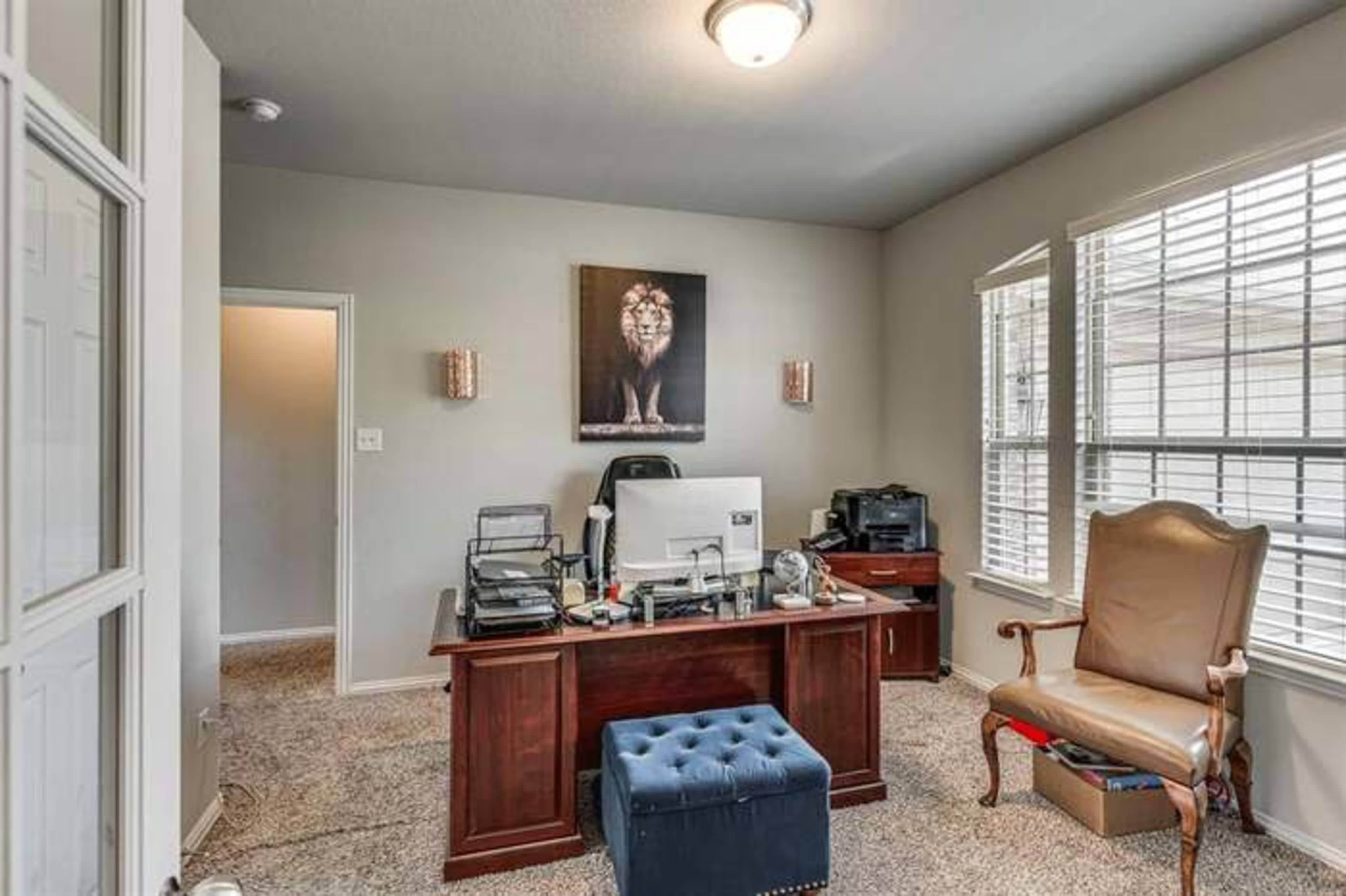 The image shows a small office with a wooden desk, a computer setup, and a chair, featuring a lion painting on the wall and a blue ottoman.