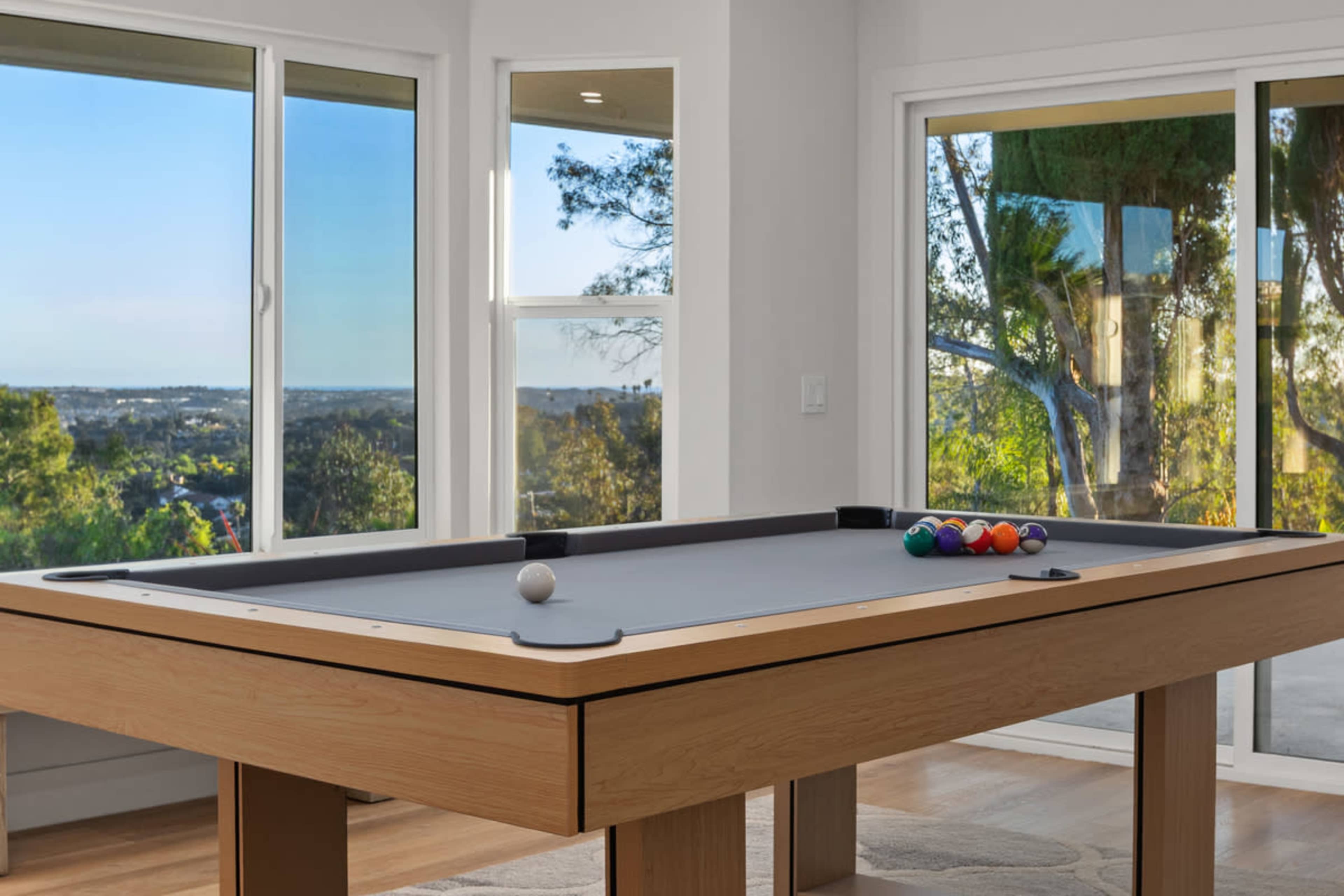 A billiards table with a set of colored balls is positioned near large windows, offering a view of a landscape.