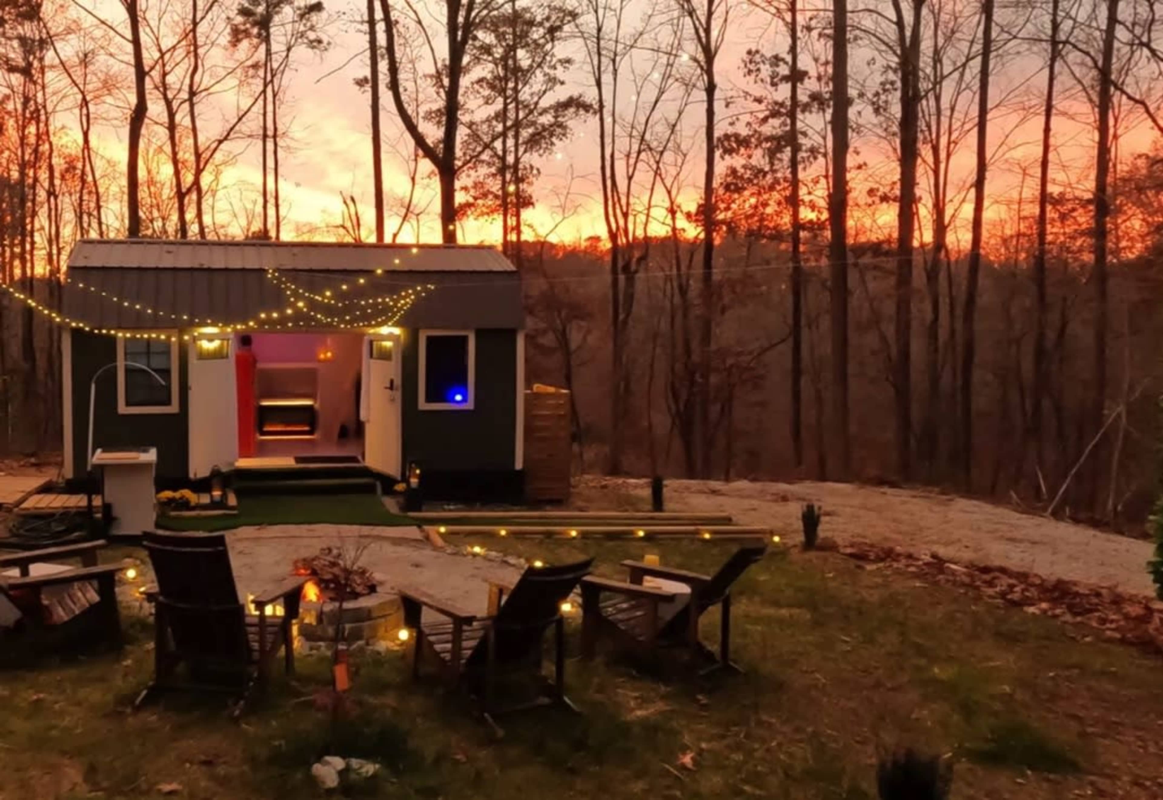 A small cabin with string lights on the porch is surrounded by outdoor seating and overlooks a forest at sunset.