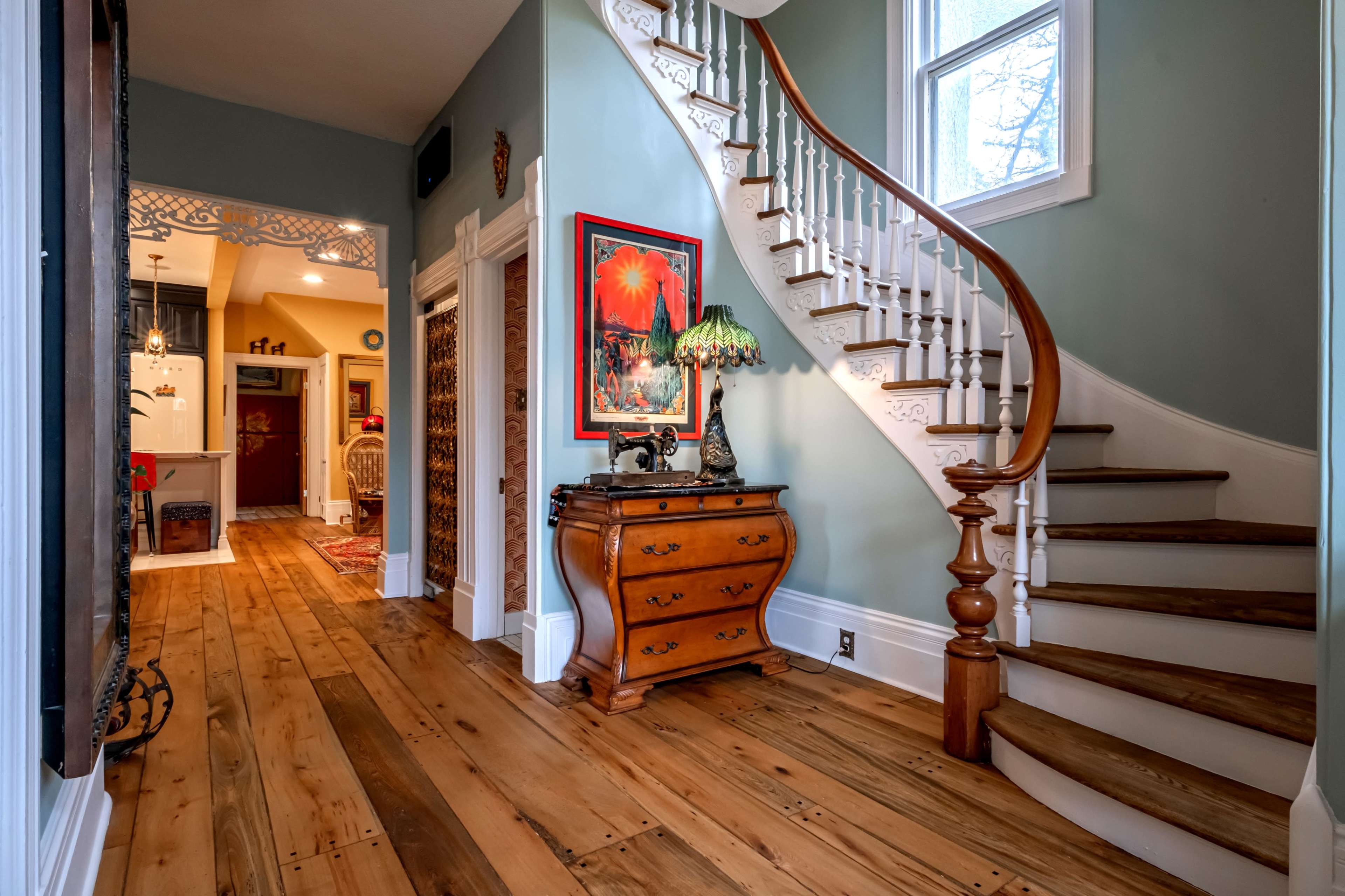 A hallway featuring a wooden staircase, a vintage chest of drawers, and a colorful artwork on the wall, with light-colored wooden flooring.
