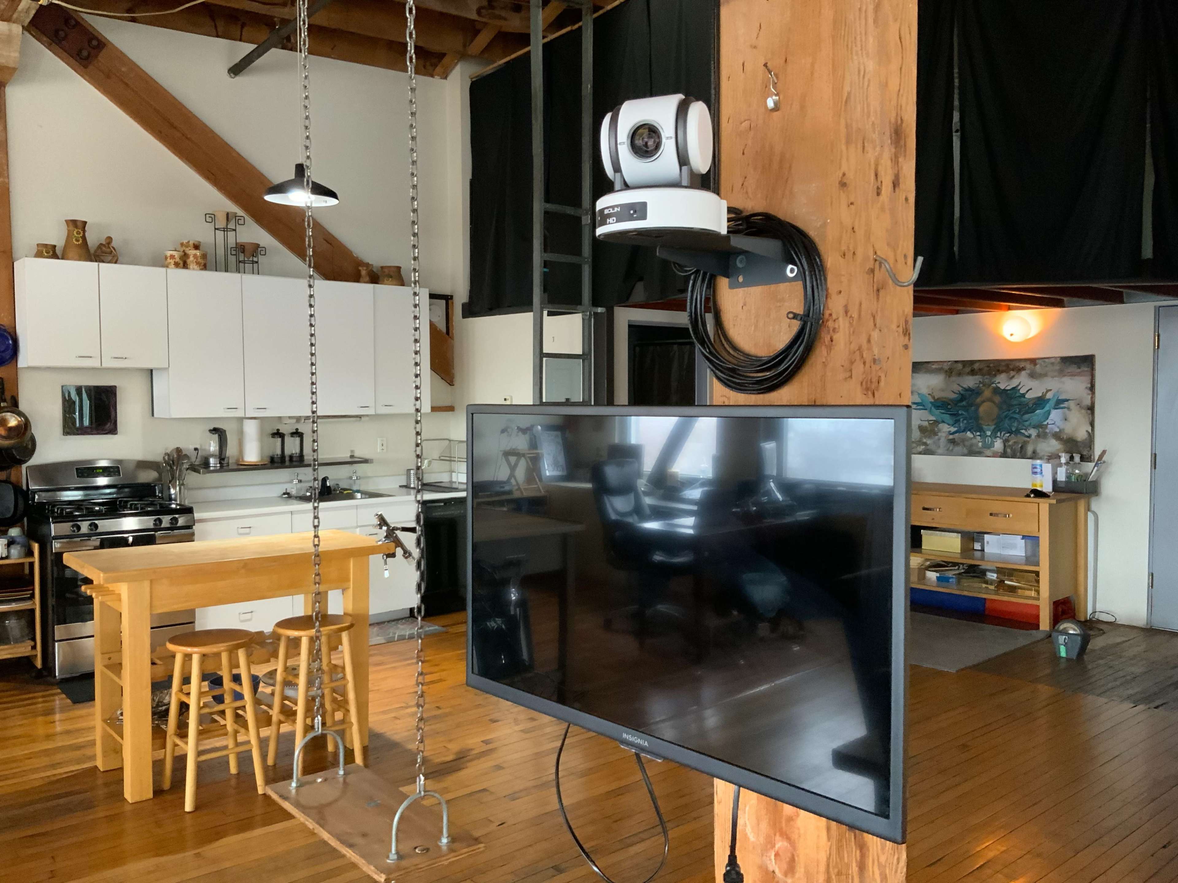 A modern kitchen and living area feature an exposed wooden beam, a mounted television, and a video camera above a dining table.