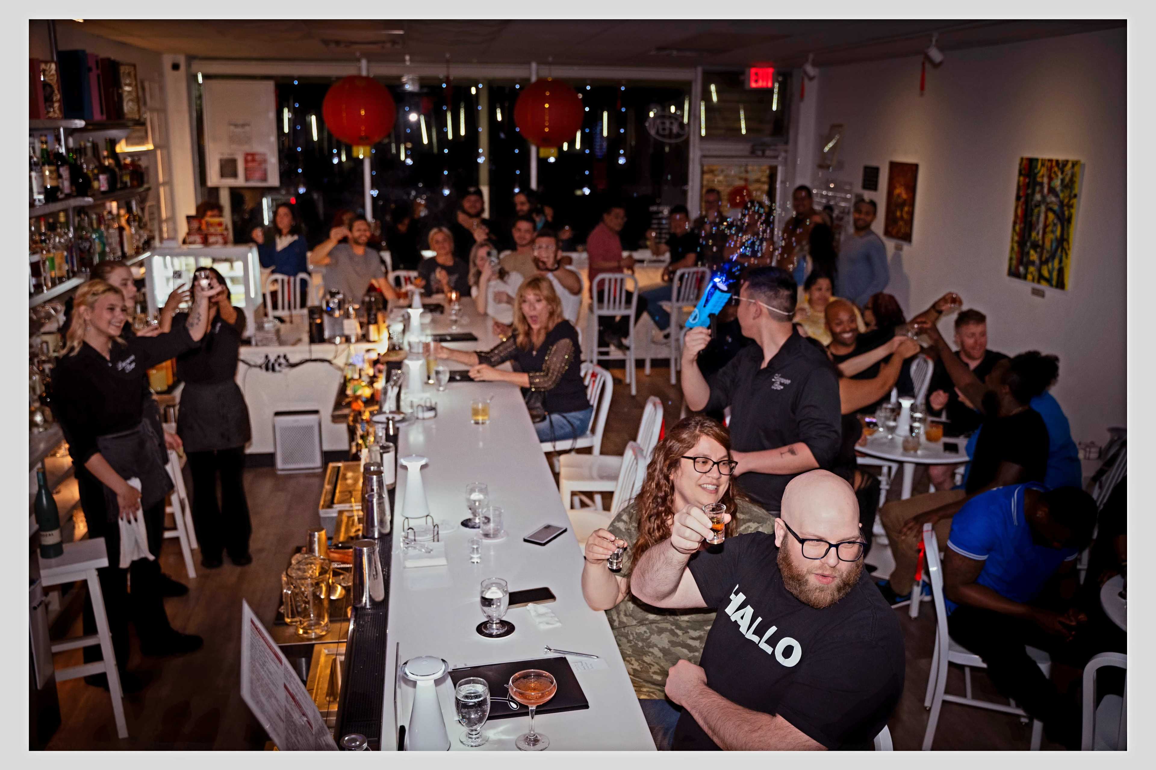 A crowded bar scene shows a mix of patrons enjoying drinks while a bartender serves at a long countertop under dim lighting and decorative elements.