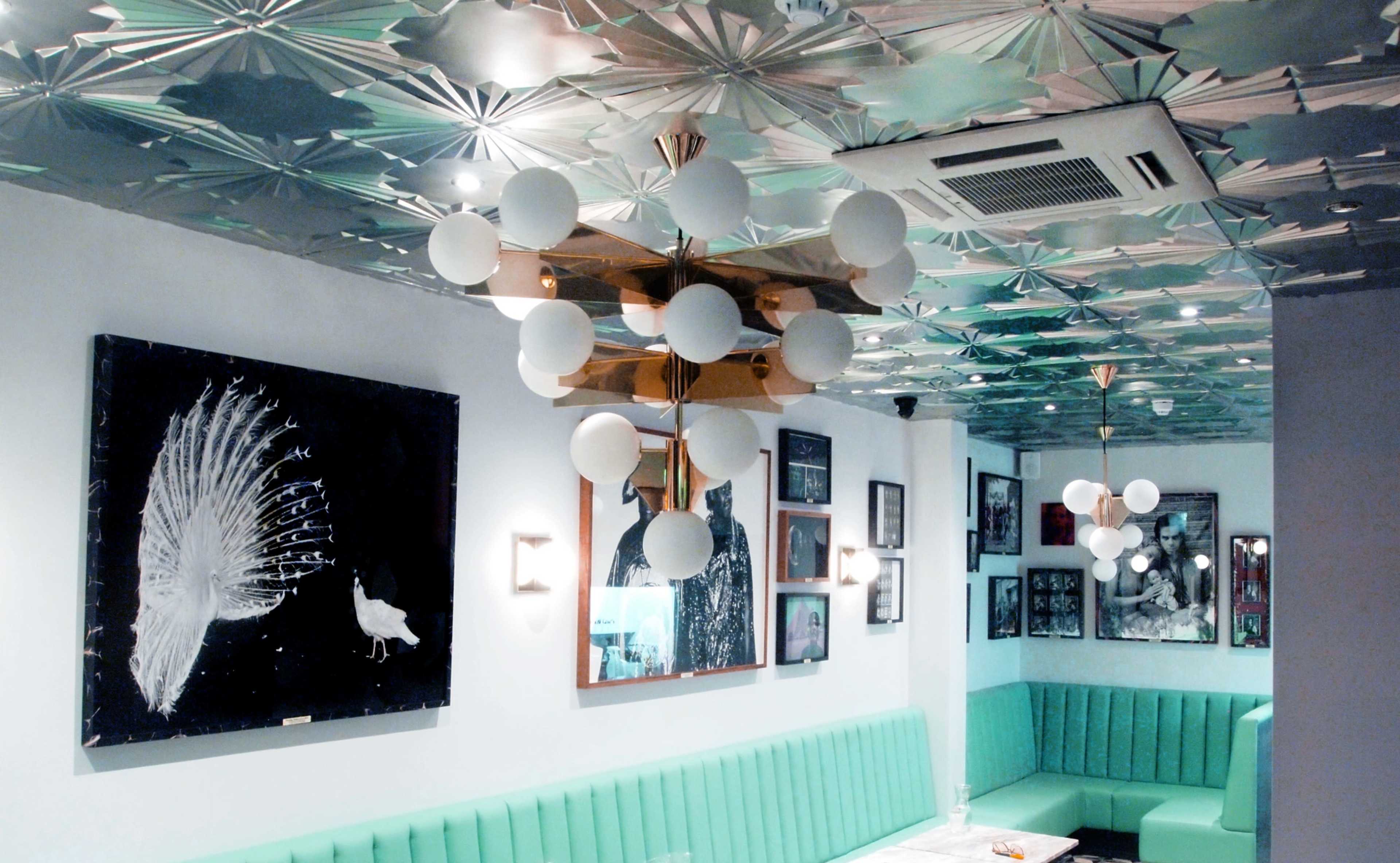 A modern restaurant interior featuring a green banquette, decorative wall art, and a distinctive ceiling with geometric patterns and pendant lighting.