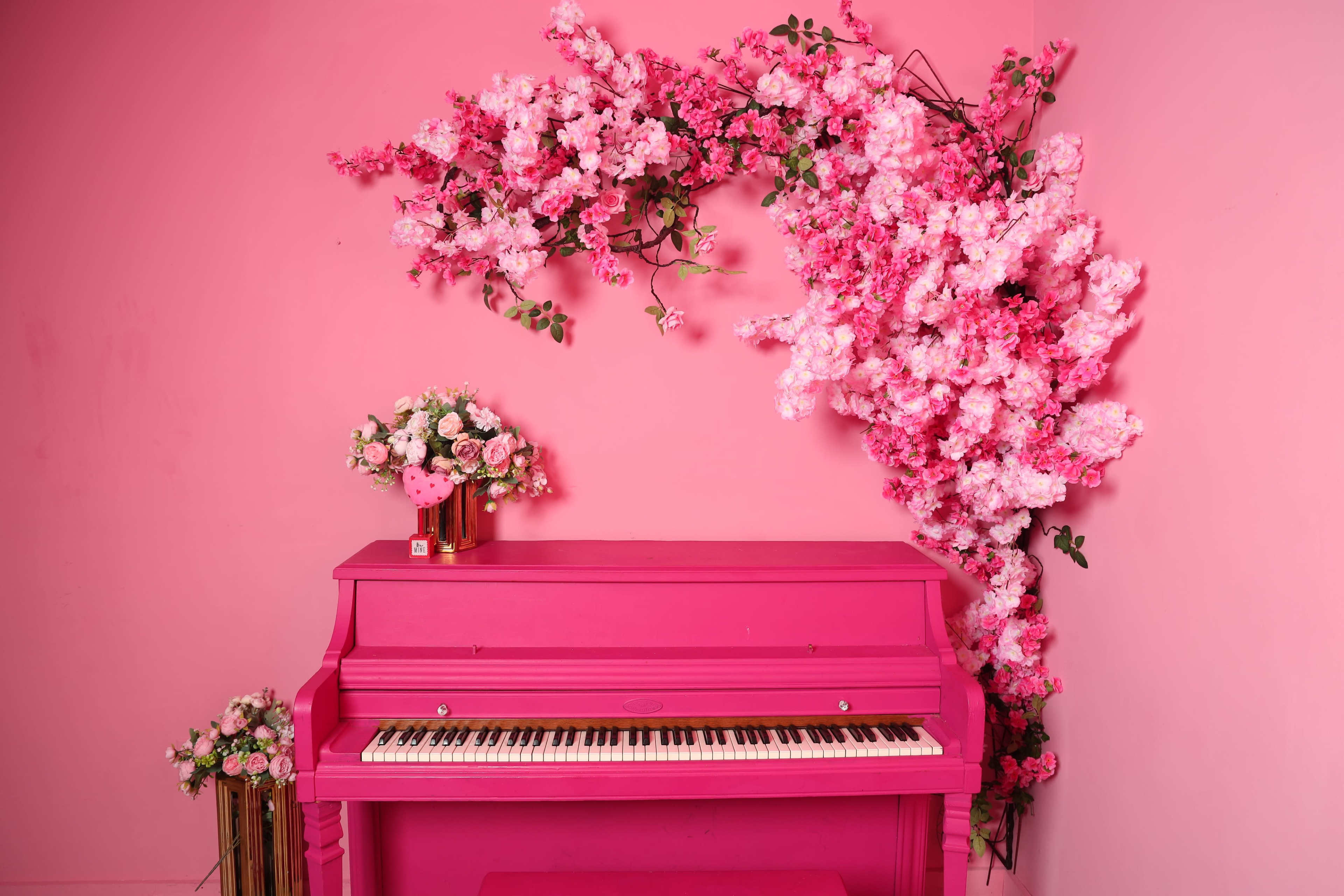 A pink piano is placed against a pink wall adorned with vibrant pink flowers and a floral arrangement on a nearby stand.