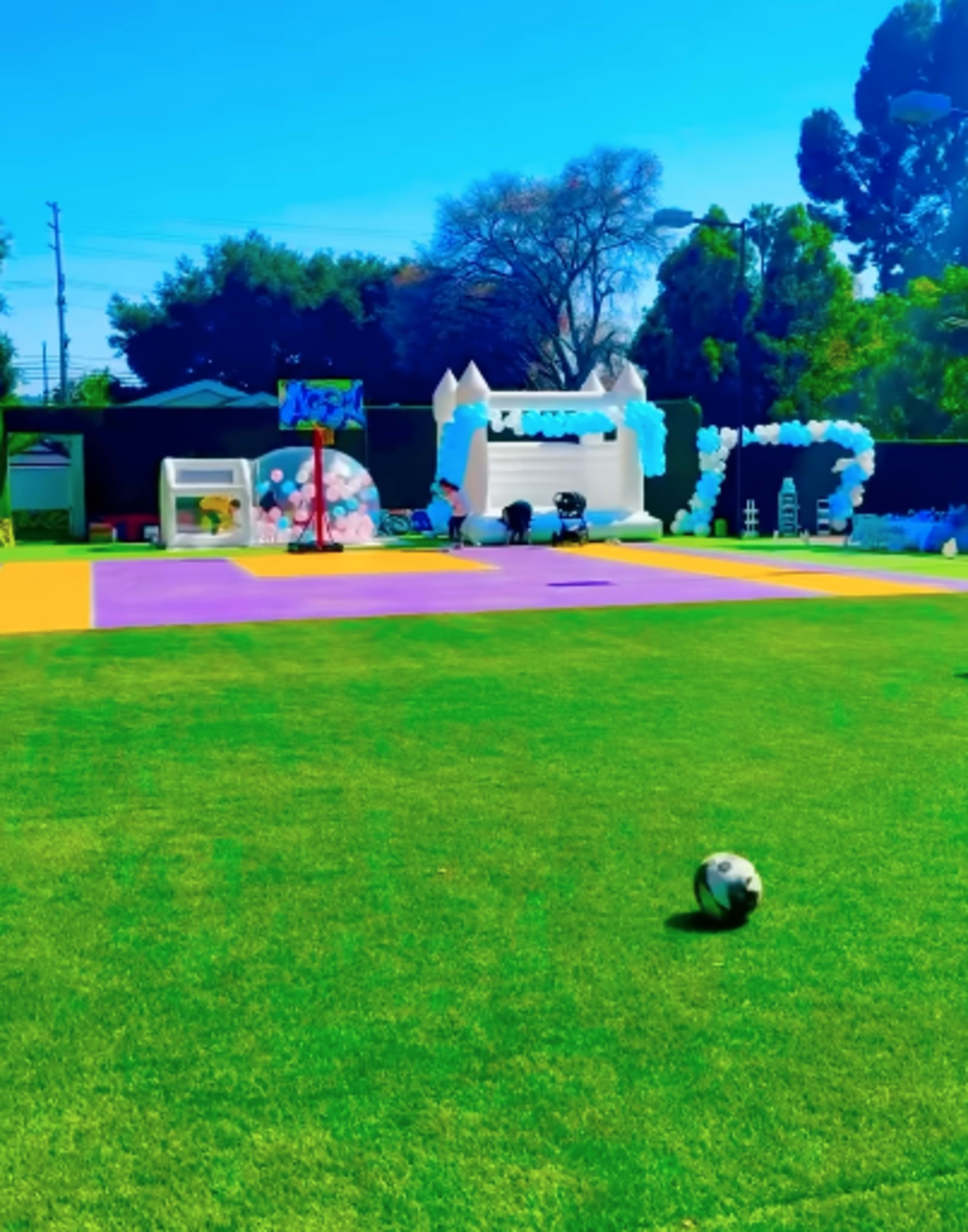 A colorful outdoor party area features a grass lawn, a soccer ball, and decorative elements including balloons and a bounce house.