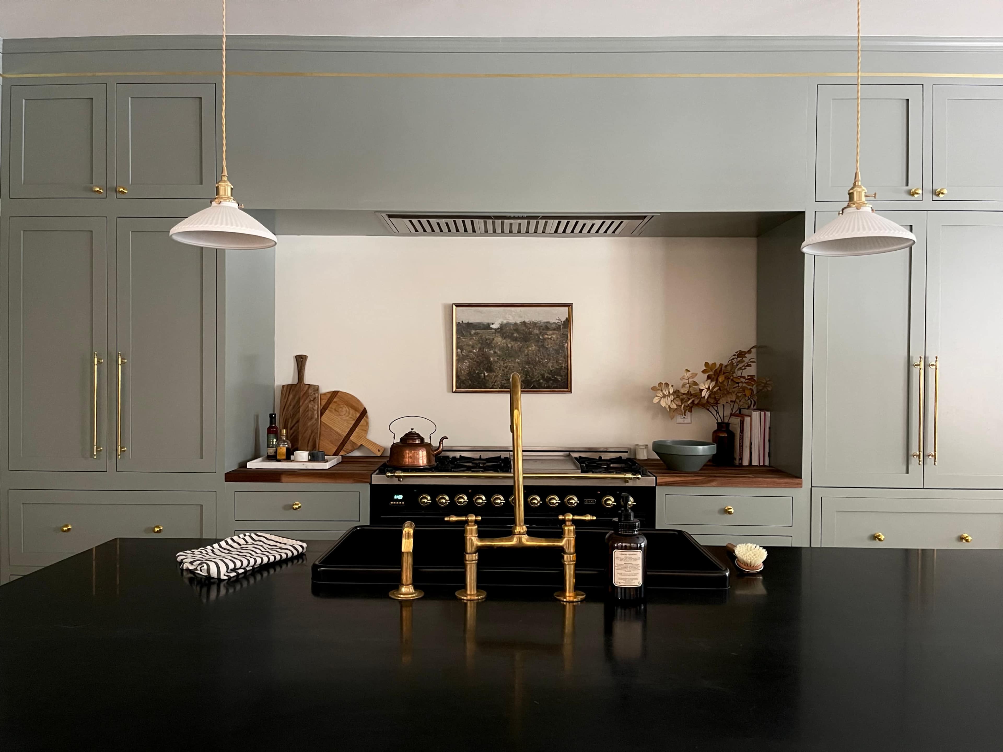 A modern kitchen features green cabinetry, a black countertop with a brass faucet, hanging light fixtures, and various kitchen utensils.