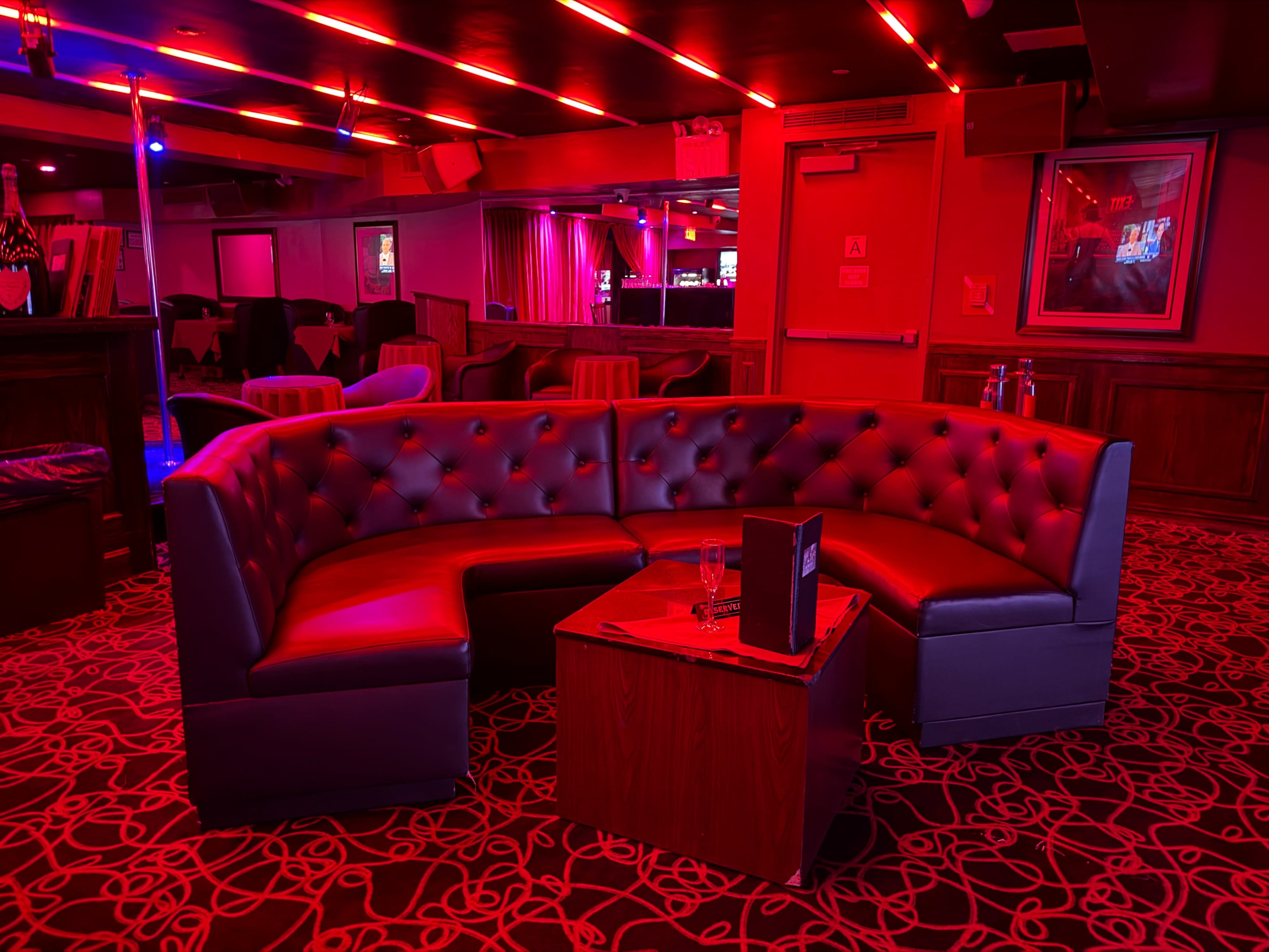 The image shows a dimly lit lounge area featuring a curved black leather sofa and a wooden table, all illuminated by red lights.