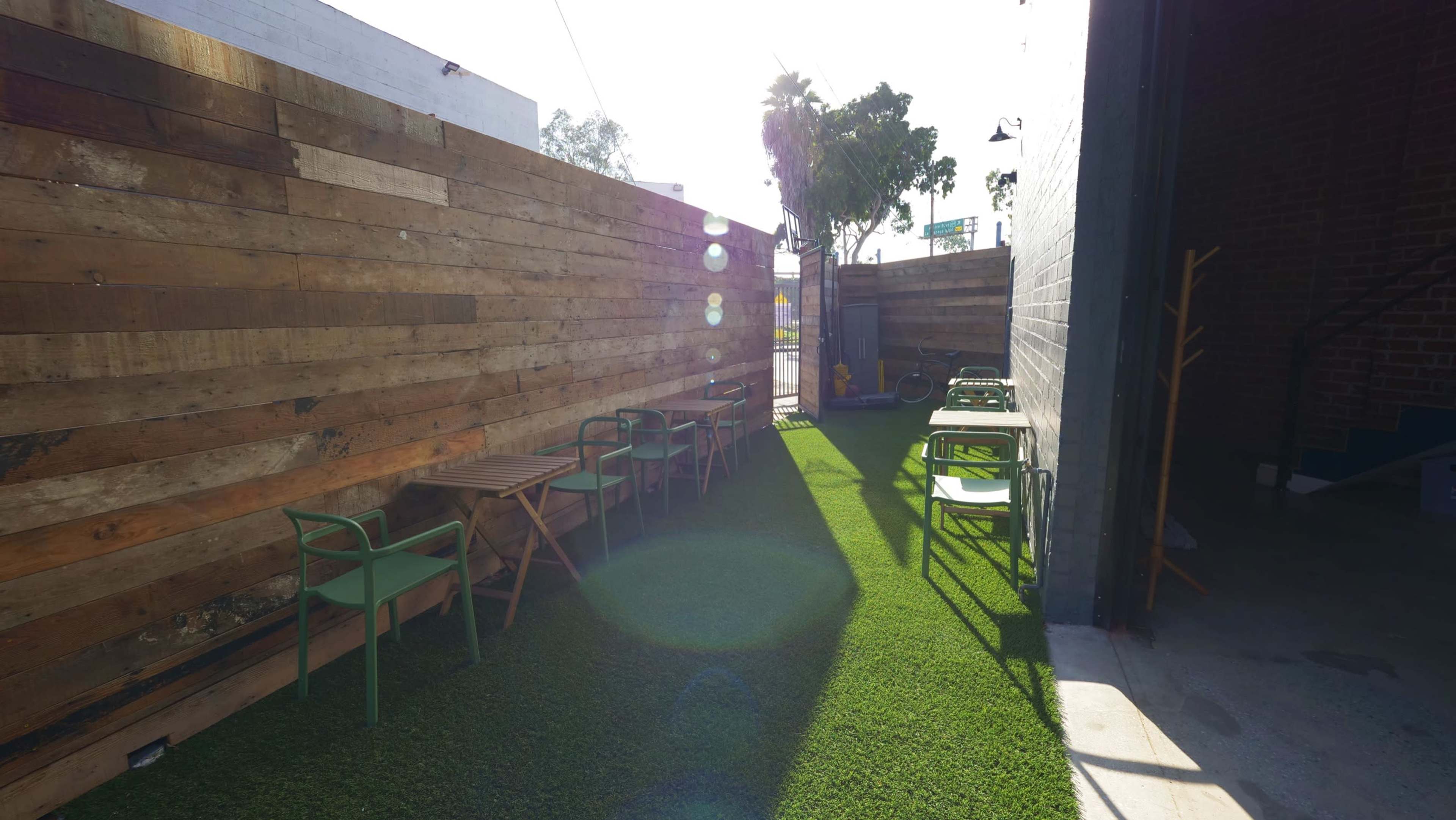 The image shows a narrow outdoor space with a wooden fence and green turf, featuring wooden and metal tables and chairs aligned along either side.