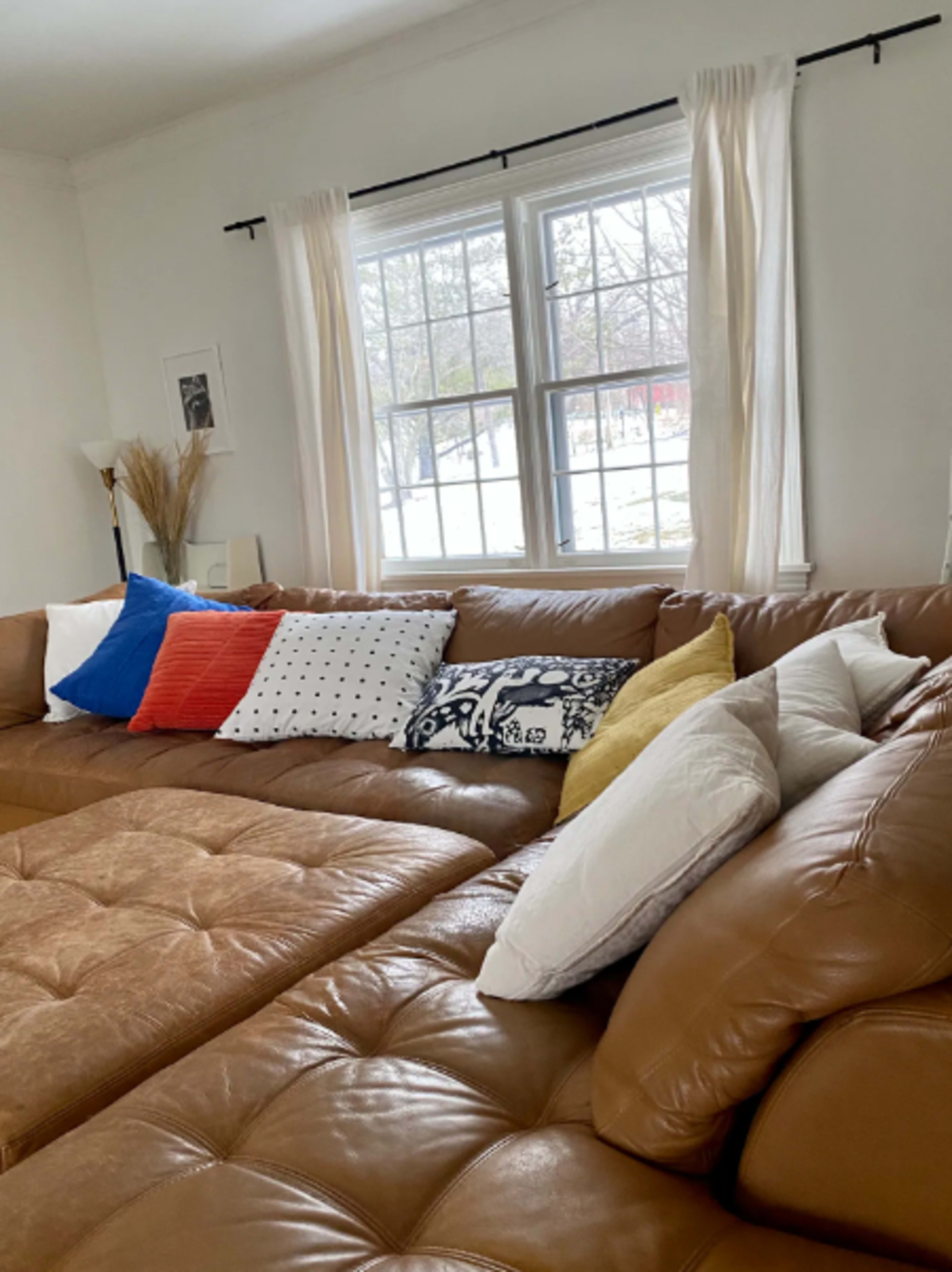 A spacious living room features a large brown sectional couch adorned with various patterned and solid color pillows, beside a window with sheer white curtains.