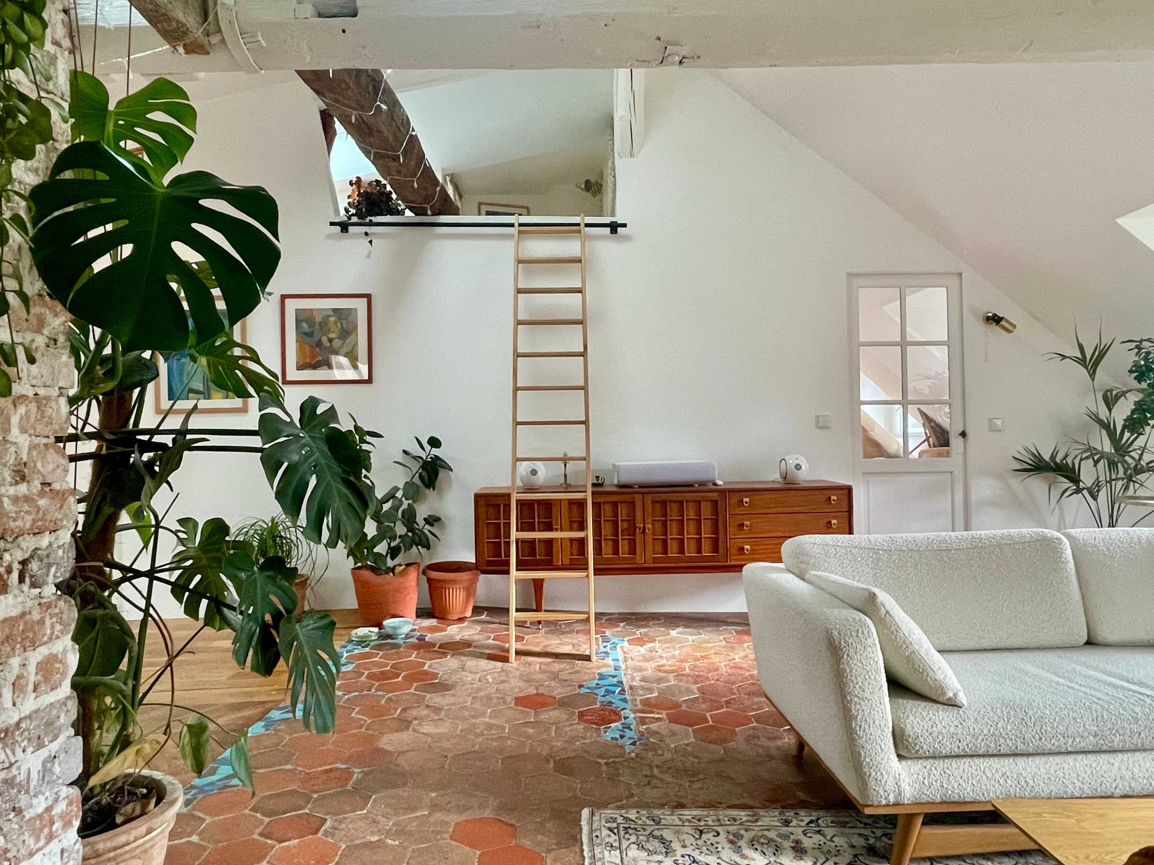 The image shows a bright, modern living area featuring a light-colored sofa, a wooden ladder leading to a loft, and various plants placed around a patterned rug.