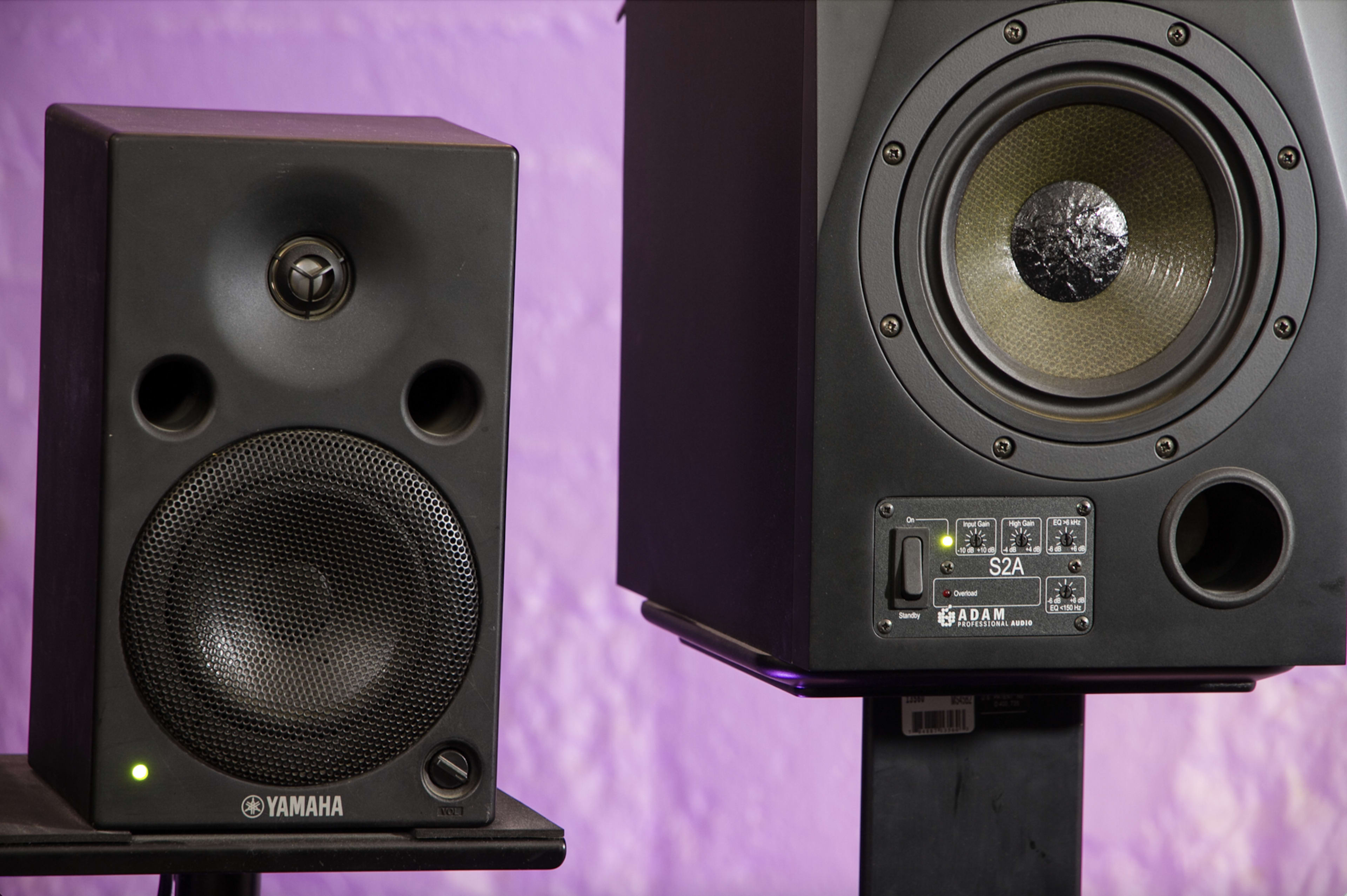 The image shows two studio monitor speakers, one branded Yamaha on the left and another branded Adam on the right, positioned against a purple wall.
