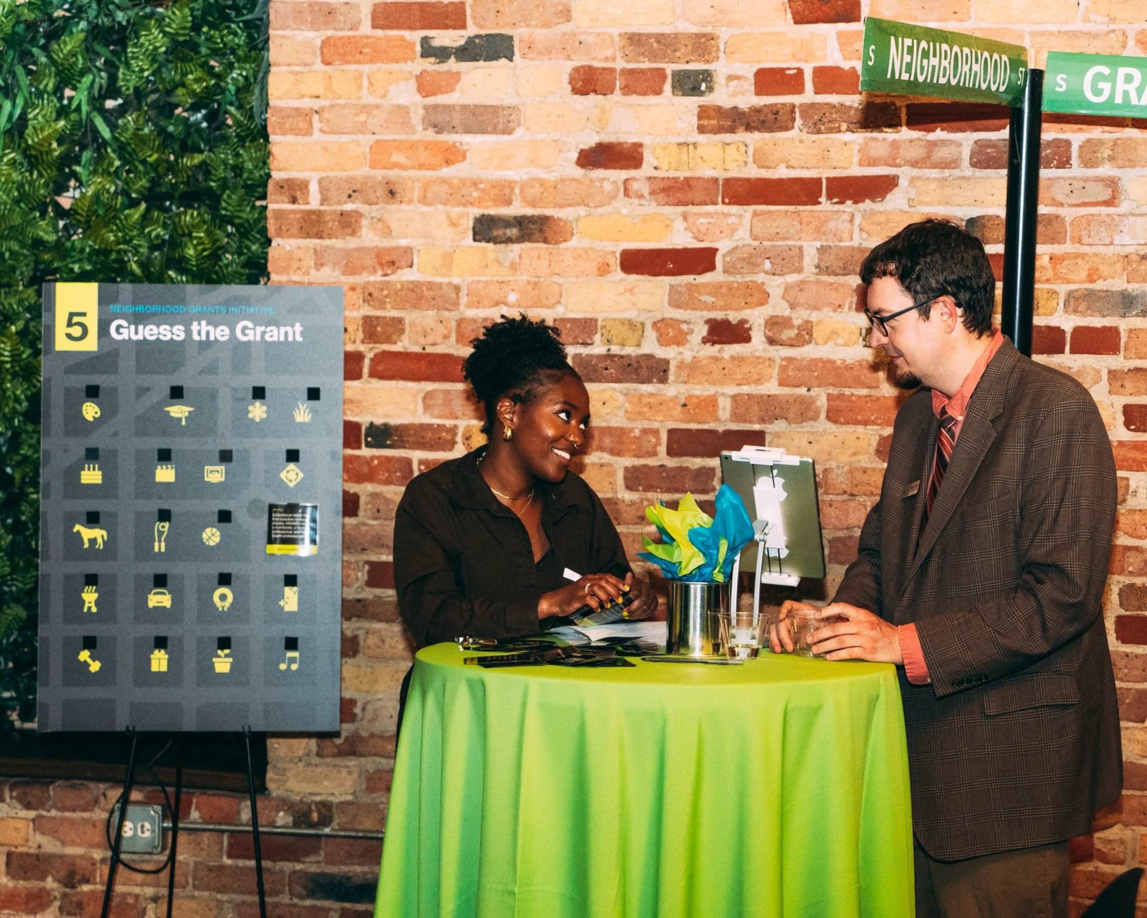 A woman and a man are engaged in conversation at a table featuring a game called "Guess the Grant" alongside a colorful display of icons related to the theme.