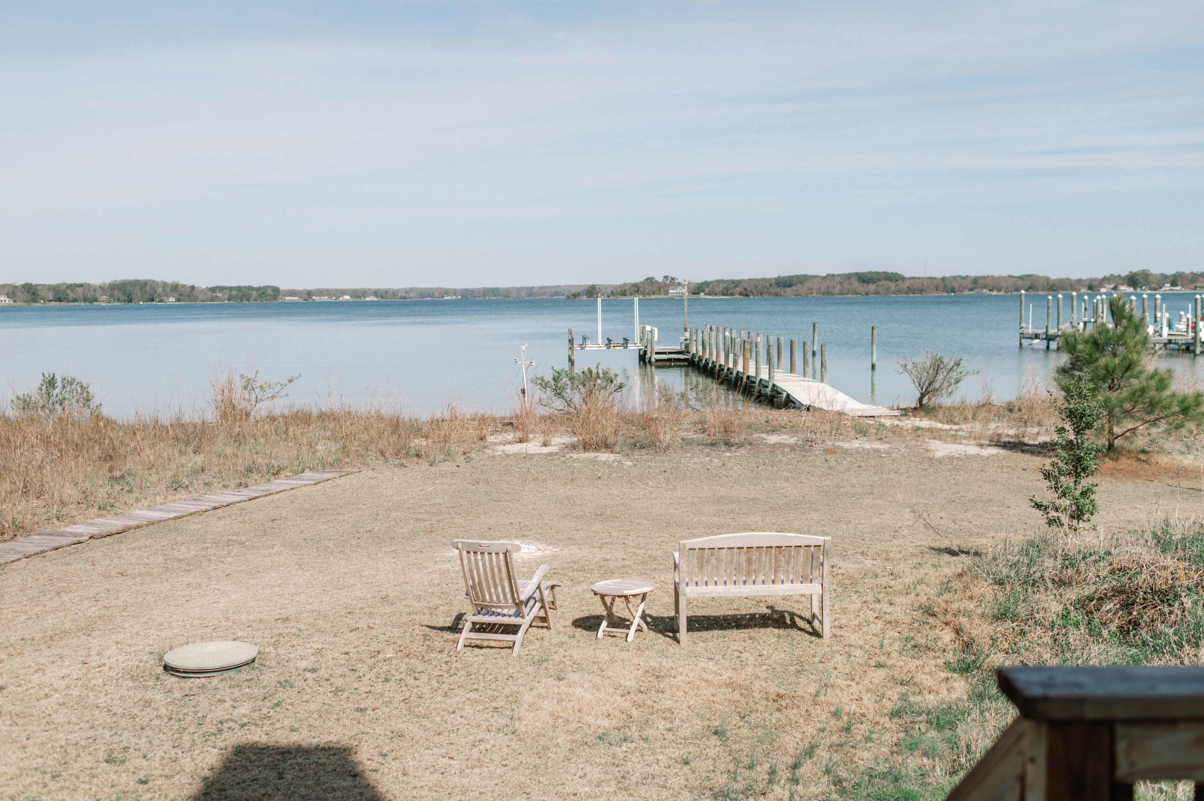 The image shows two wooden chairs and a small round table on a grass area overlooking a calm body of water and a dock.