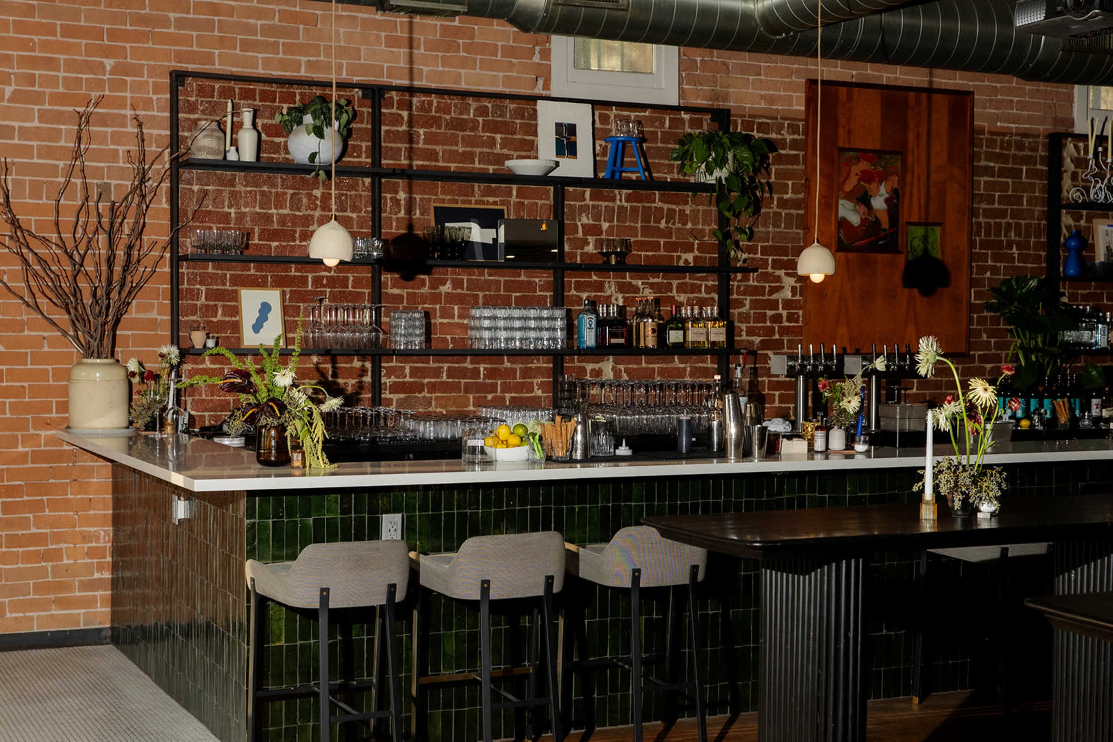 The image shows a modern bar area with a brick wall, shelves displaying various bottles, and several bar stools arranged in front of a long countertop.