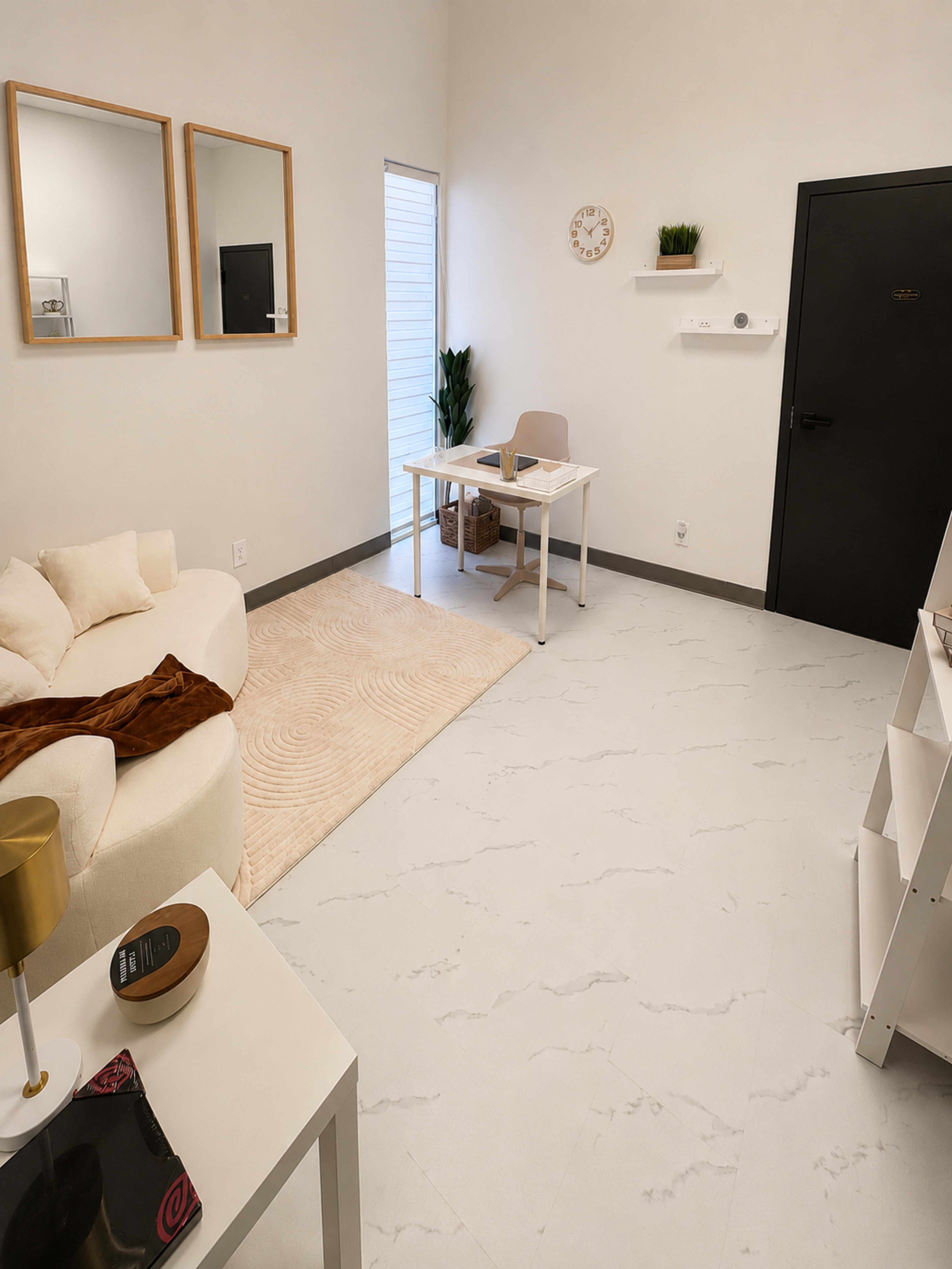 The image shows a minimalist room featuring a white sofa, a desk with a chair, and a wall-mounted clock, with light-colored flooring and a potted plant in the corner.