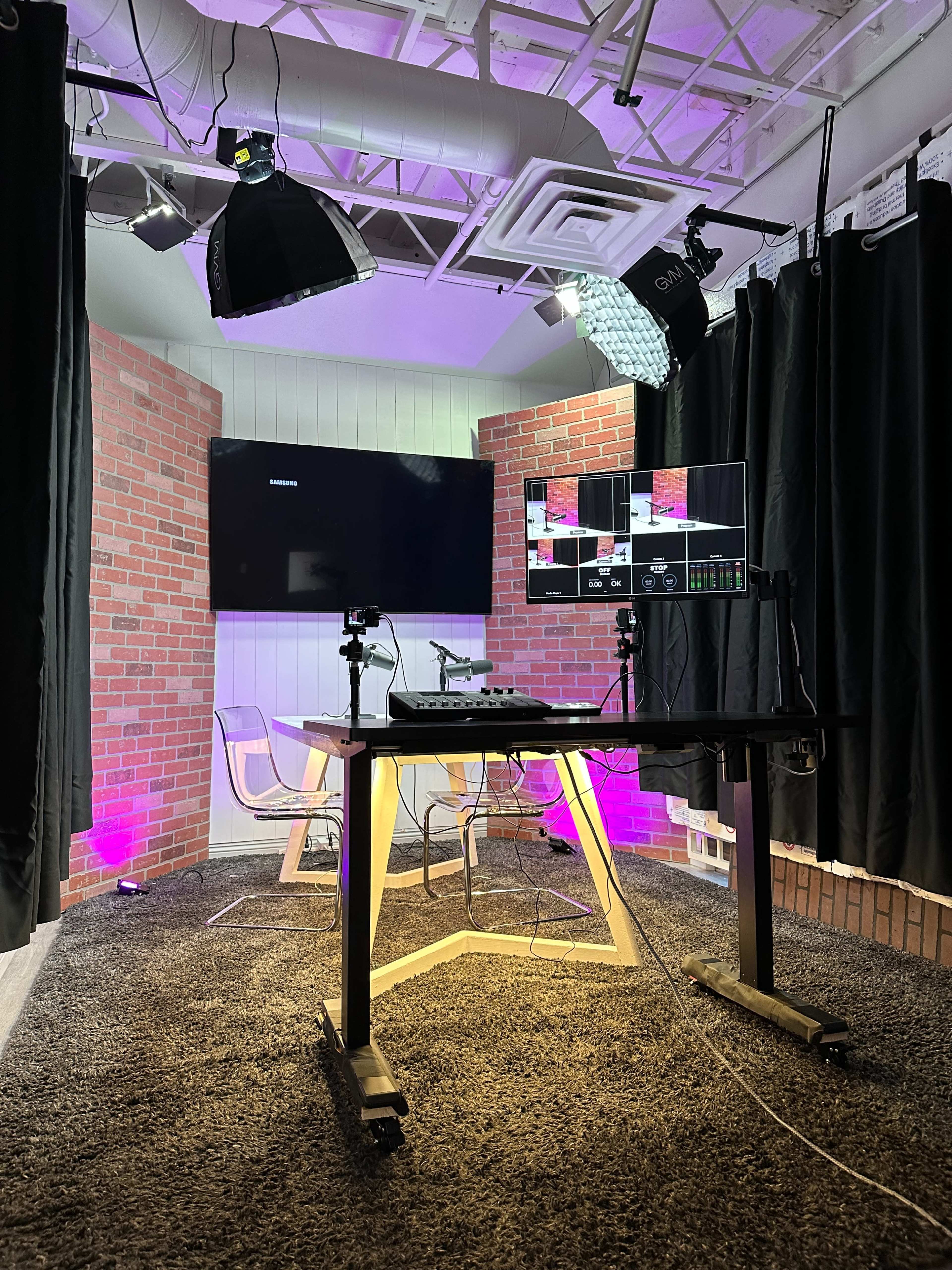 A video production studio with a desk, multiple screens, and brick wall accents, illuminated by soft lighting.