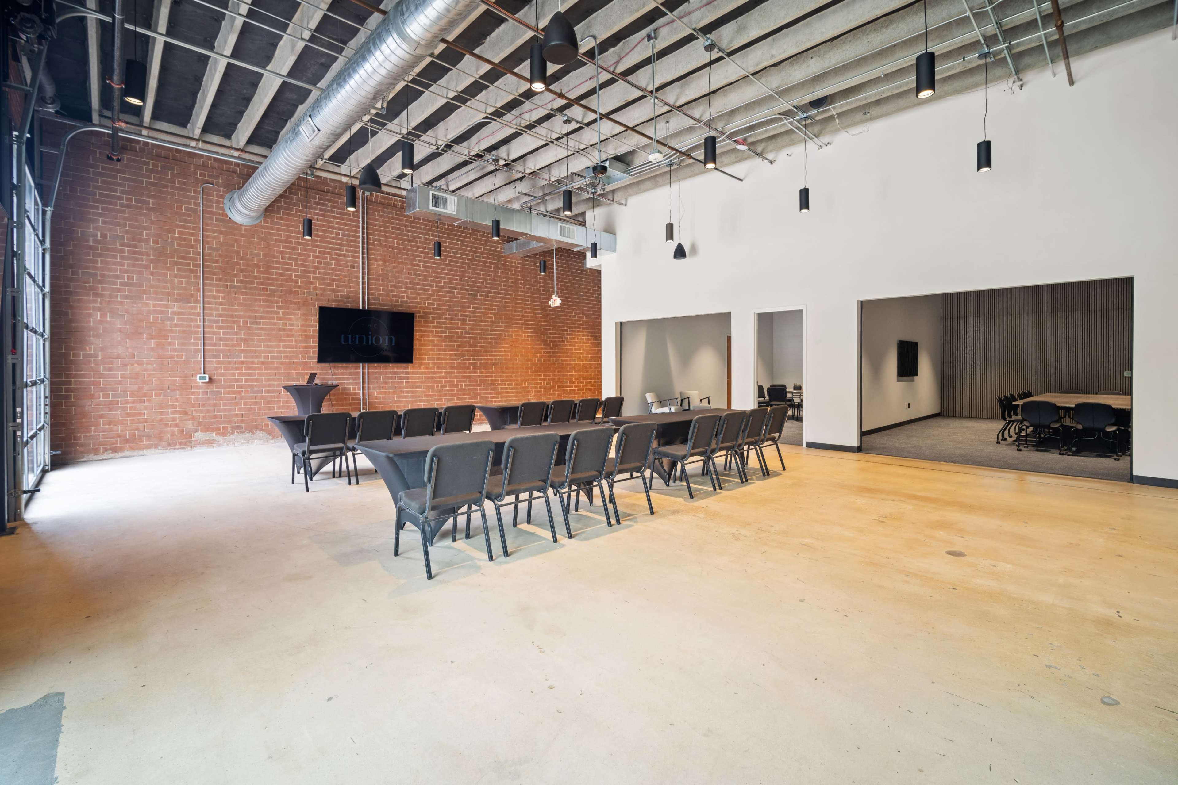 The image shows a spacious conference room featuring a long table set with chairs, exposed brick walls, and industrial-style lighting.