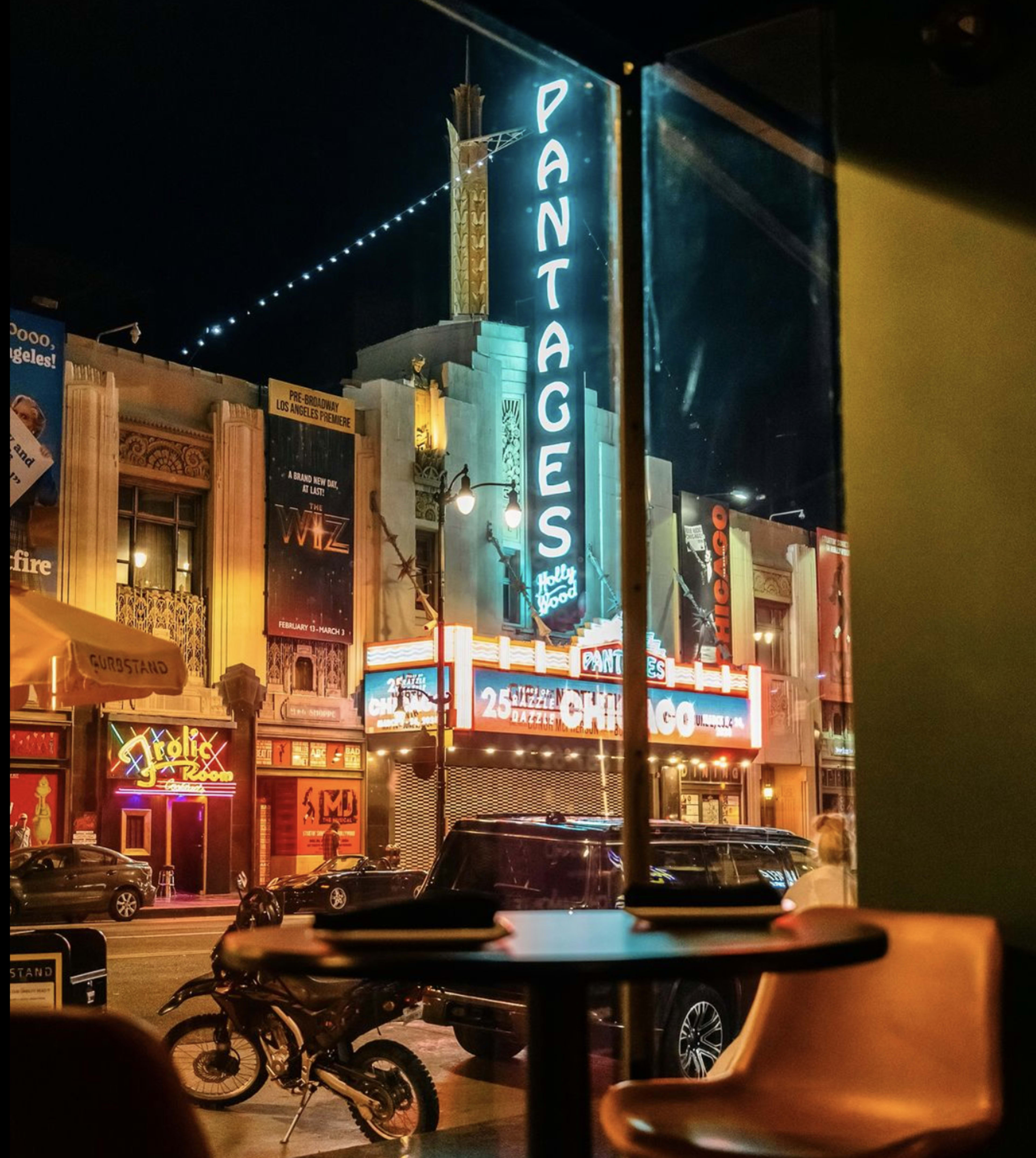 A nighttime view of the Pantages Theatre with illuminated signage and surrounding venues, seen through a window from an interior setting.
