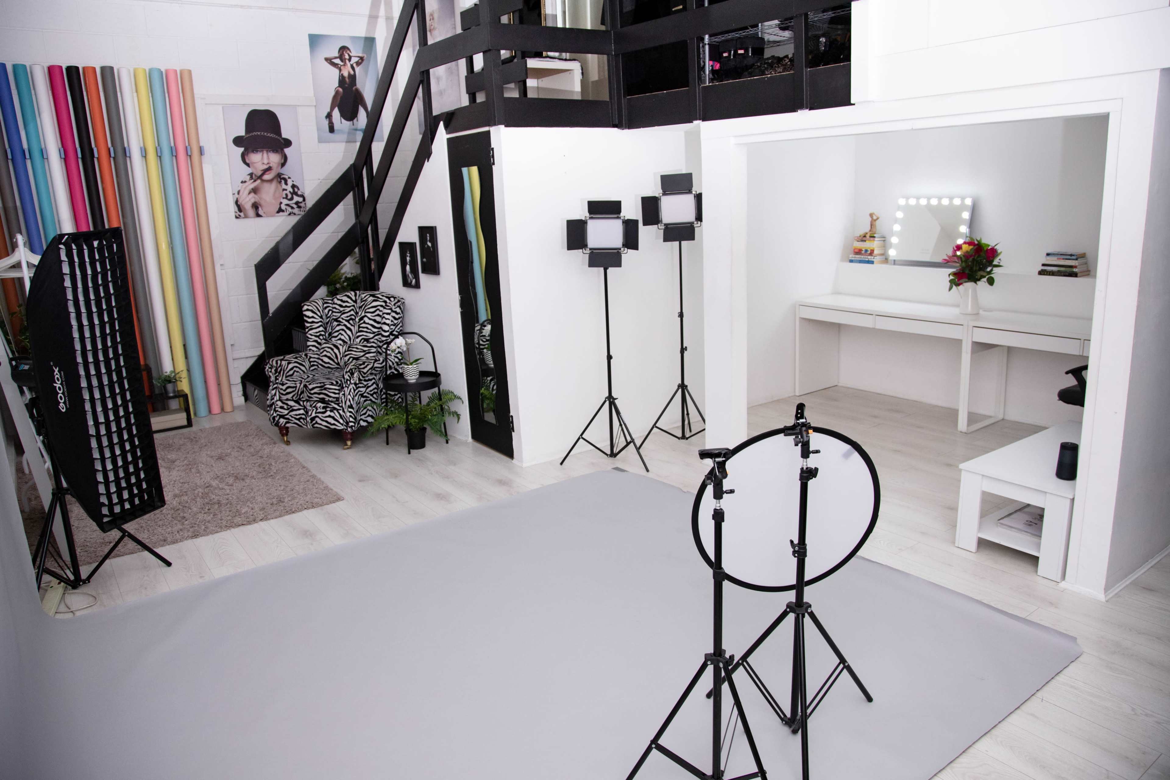 The image depicts a photography studio featuring a gray backdrop, lighting equipment, and a dressed vanity area with a mirror and flowers.