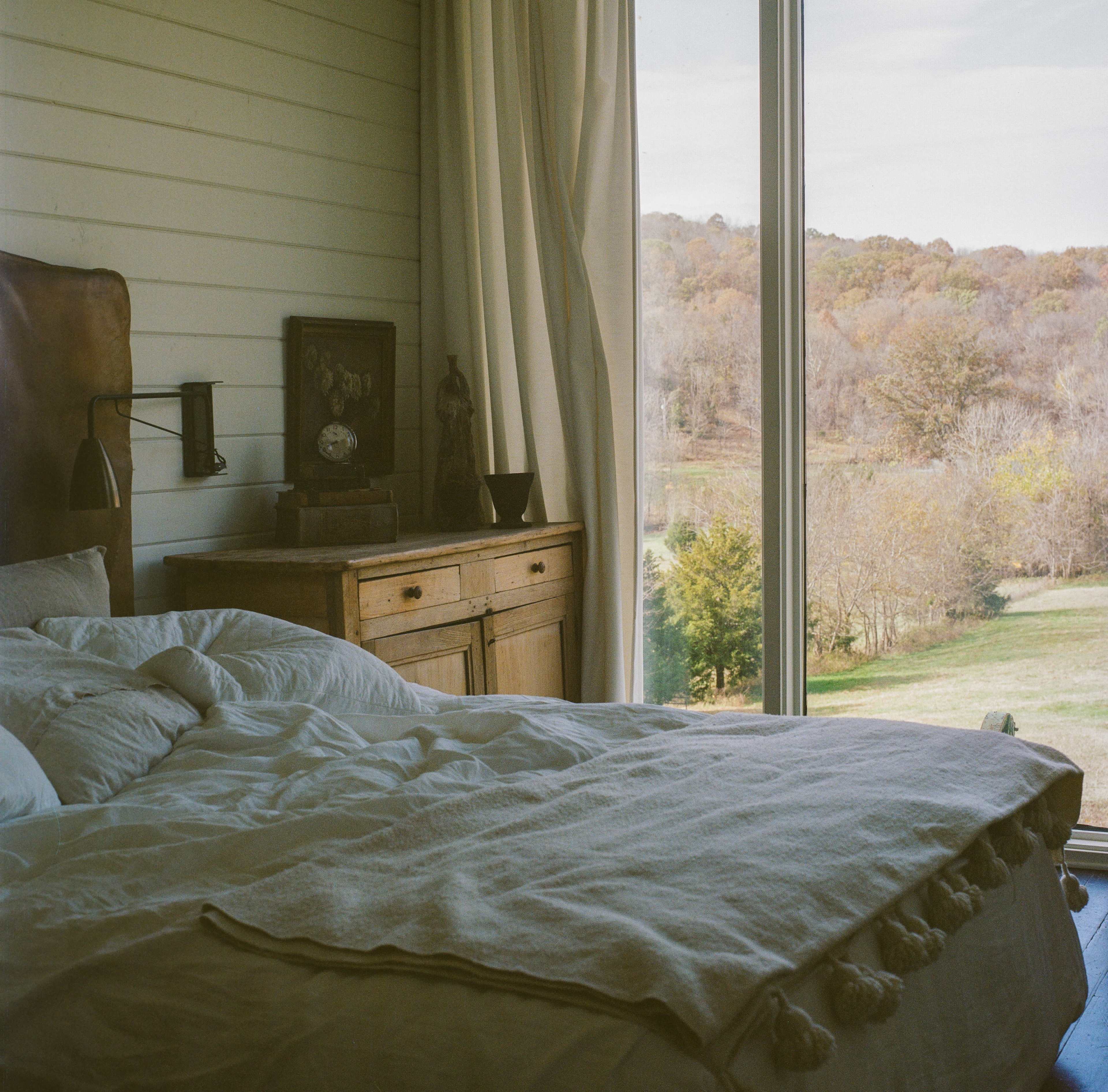 A neatly made bed with a light-colored blanket is positioned in front of a large window overlooking a landscape of trees and hills.