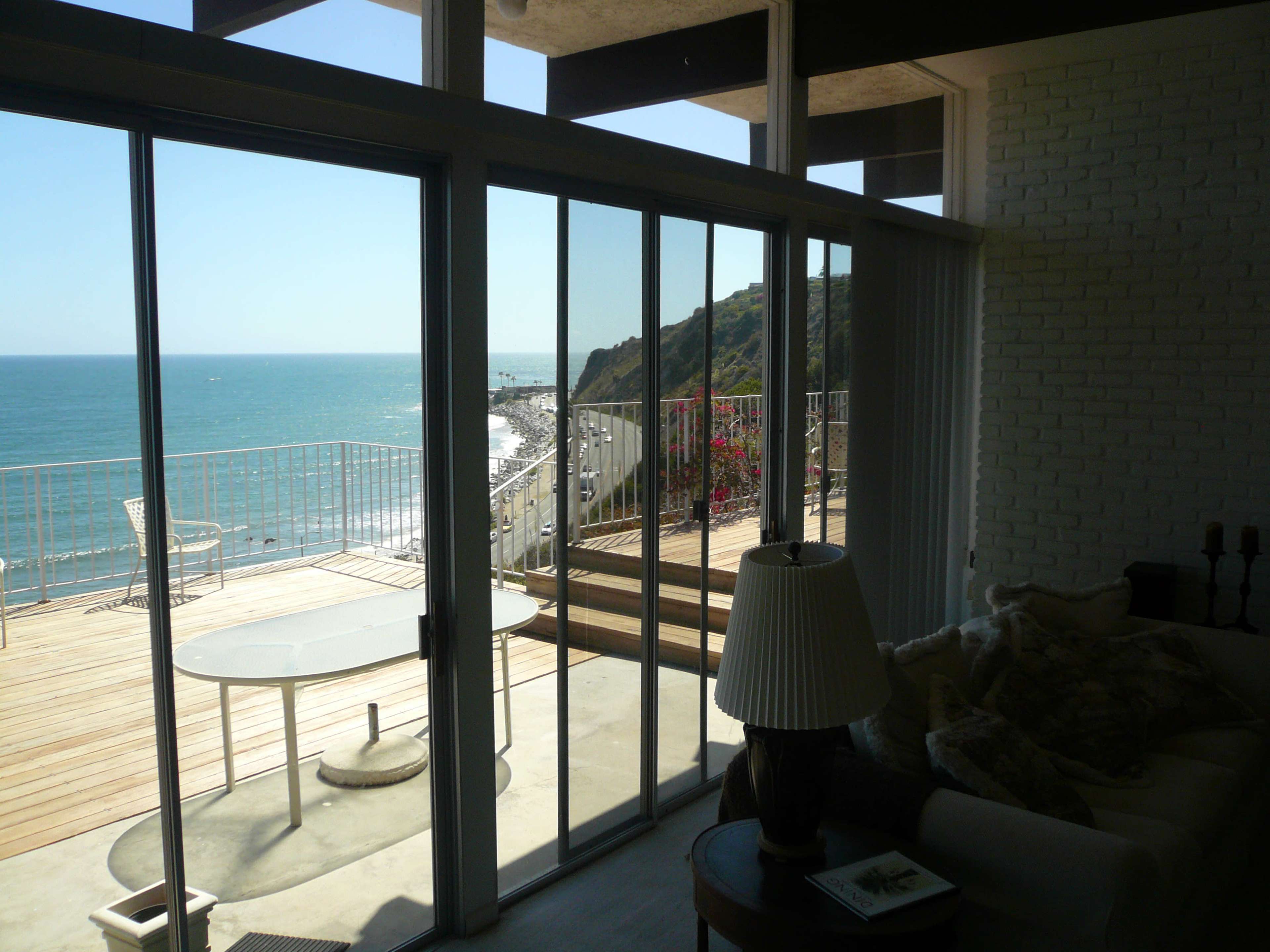 The image shows a view from an interior space with sliding glass doors open to a deck overlooking the ocean and a coastal landscape.
