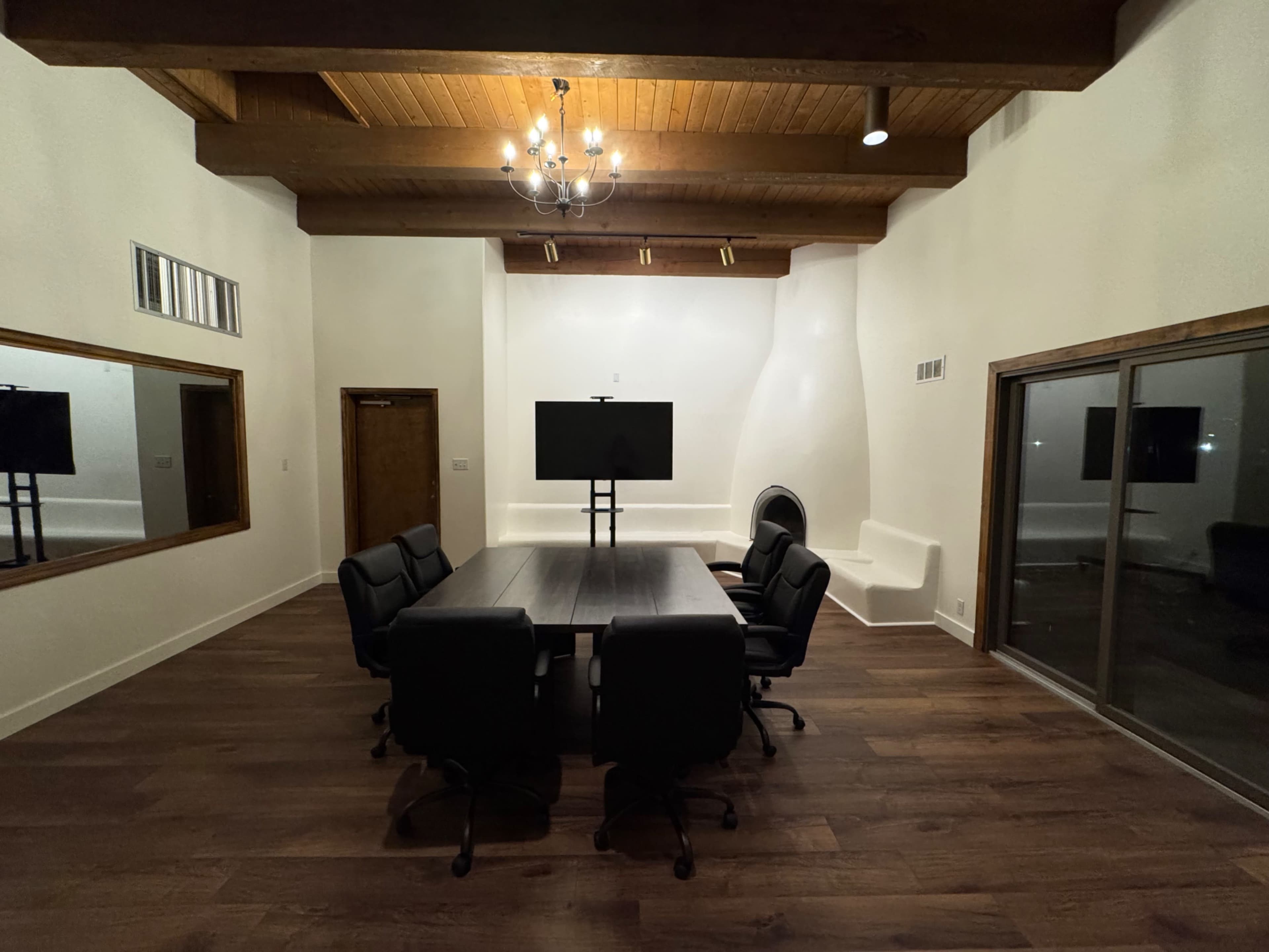 A conference room is furnished with a large table, black chairs, and a wall-mounted television, featuring wooden beams on the ceiling and a polished floor.