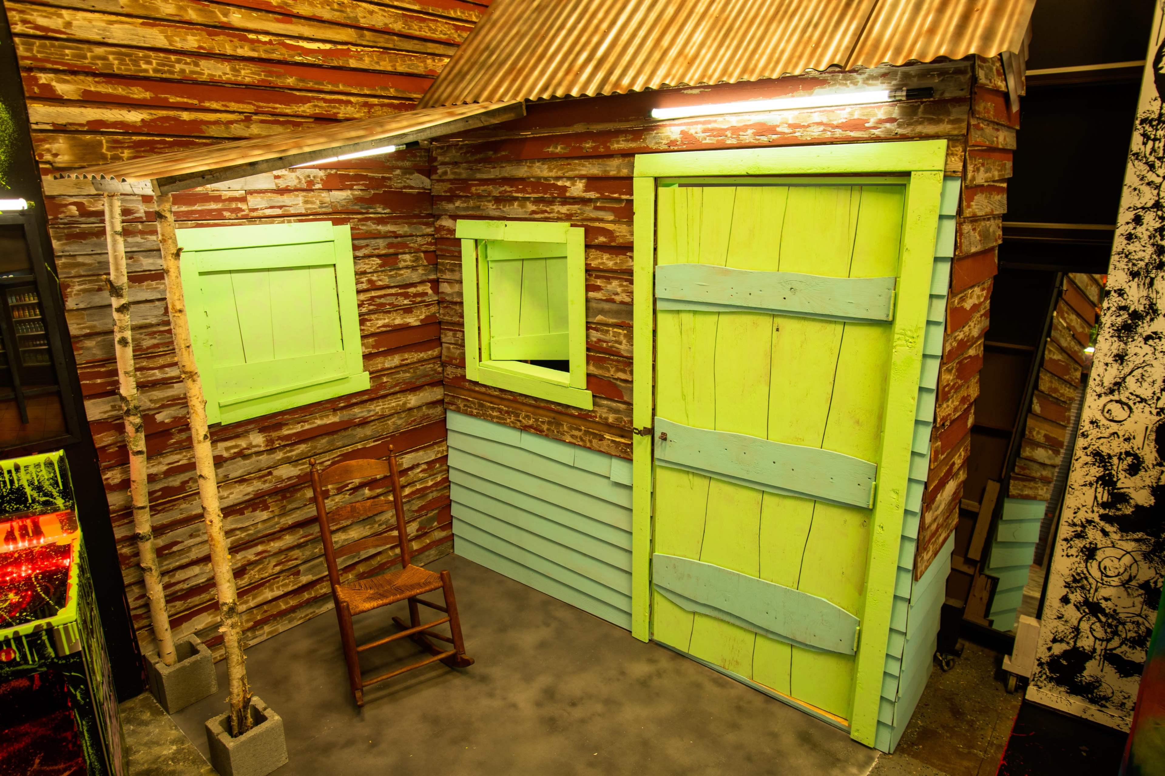 The image shows a small, rustic wooden shed with a green door and windows, featuring a corrugated metal roof and a wooden chair beside it.