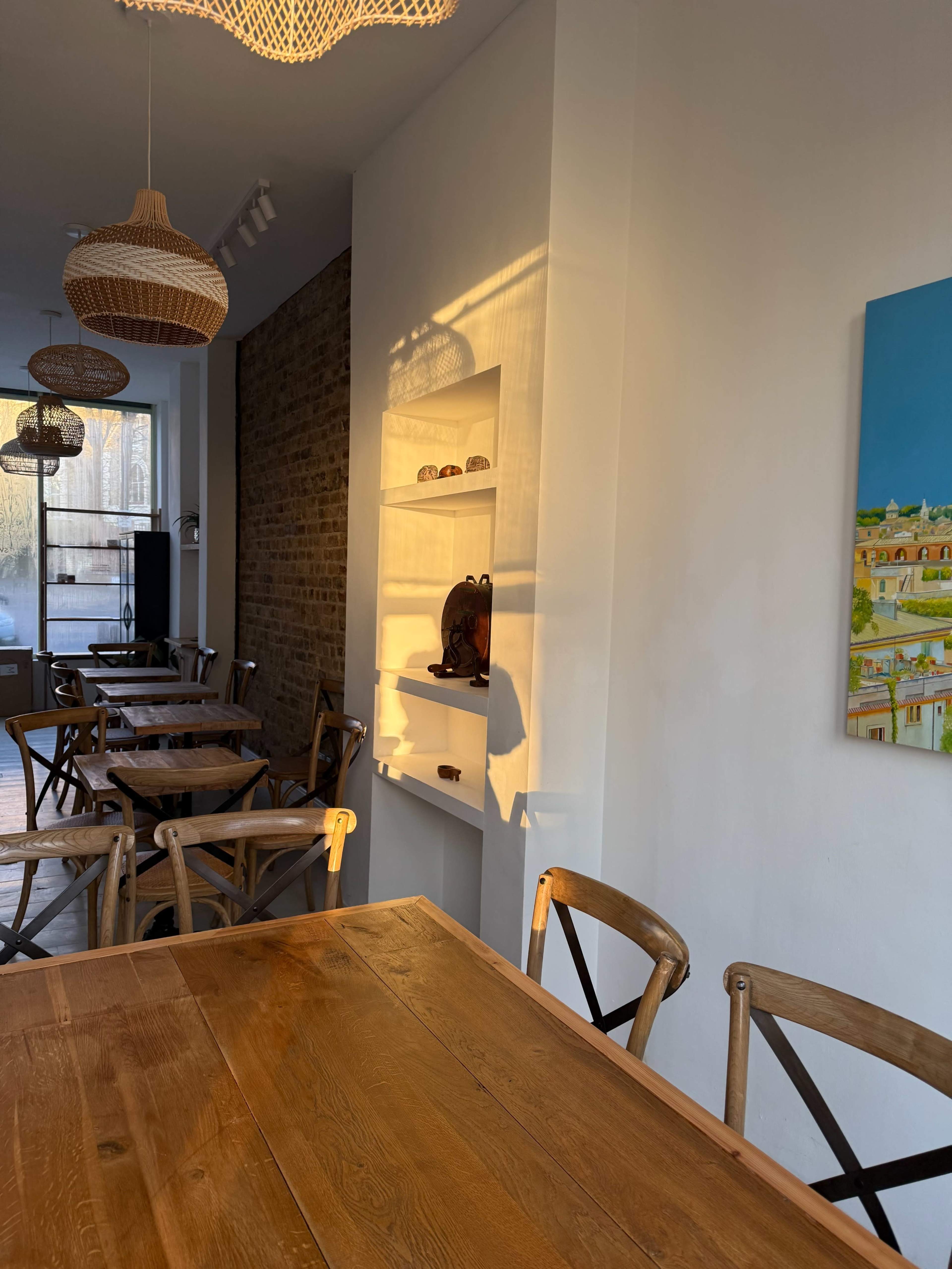 The image shows a brightly lit café interior with wooden tables, woven light fixtures, and a white shelf displaying decor items against a brick wall.