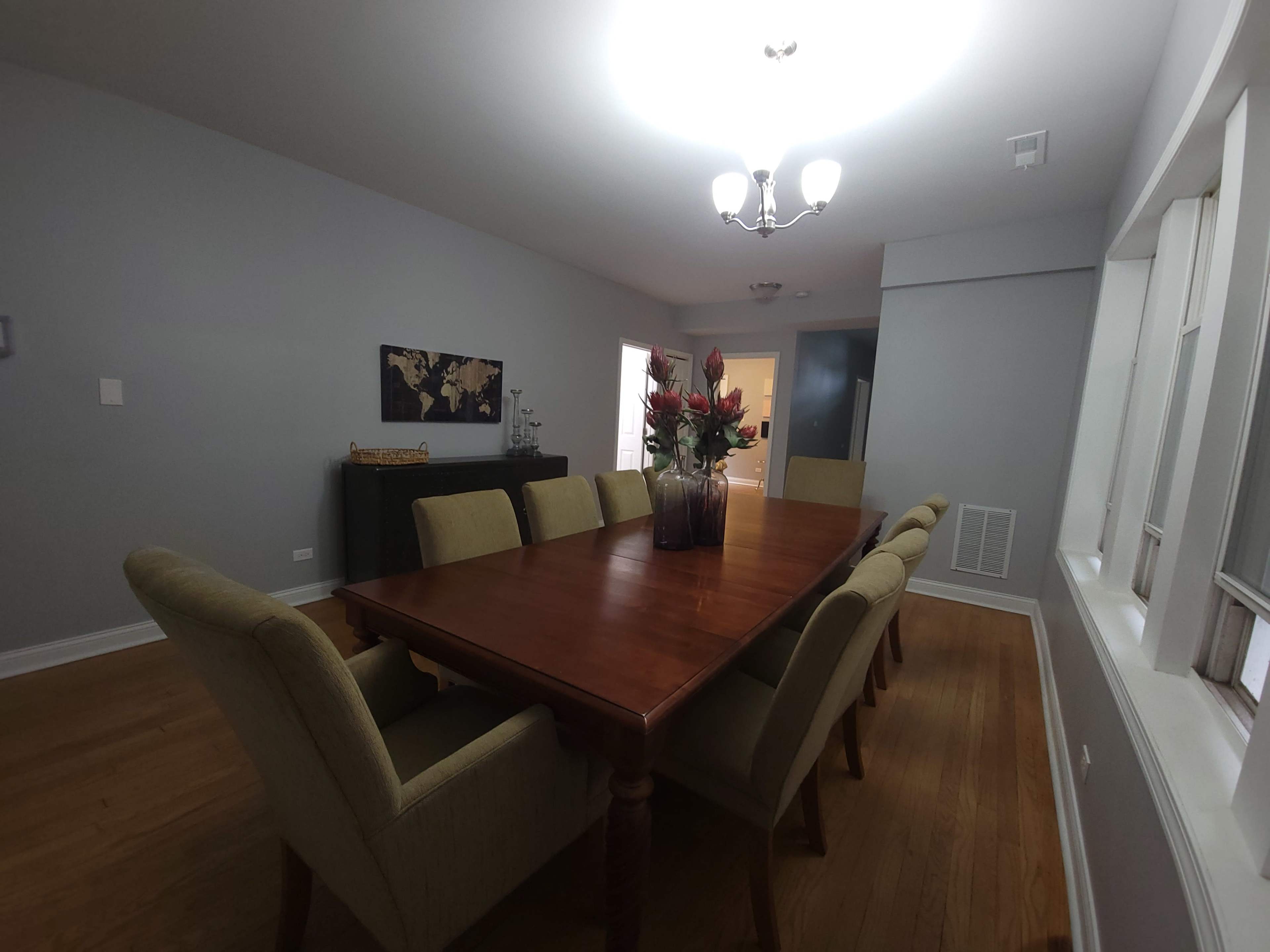 A long wooden dining table with eight chairs is centered in a bright dining room featuring a world map on the wall and a vase of flowers on the table.