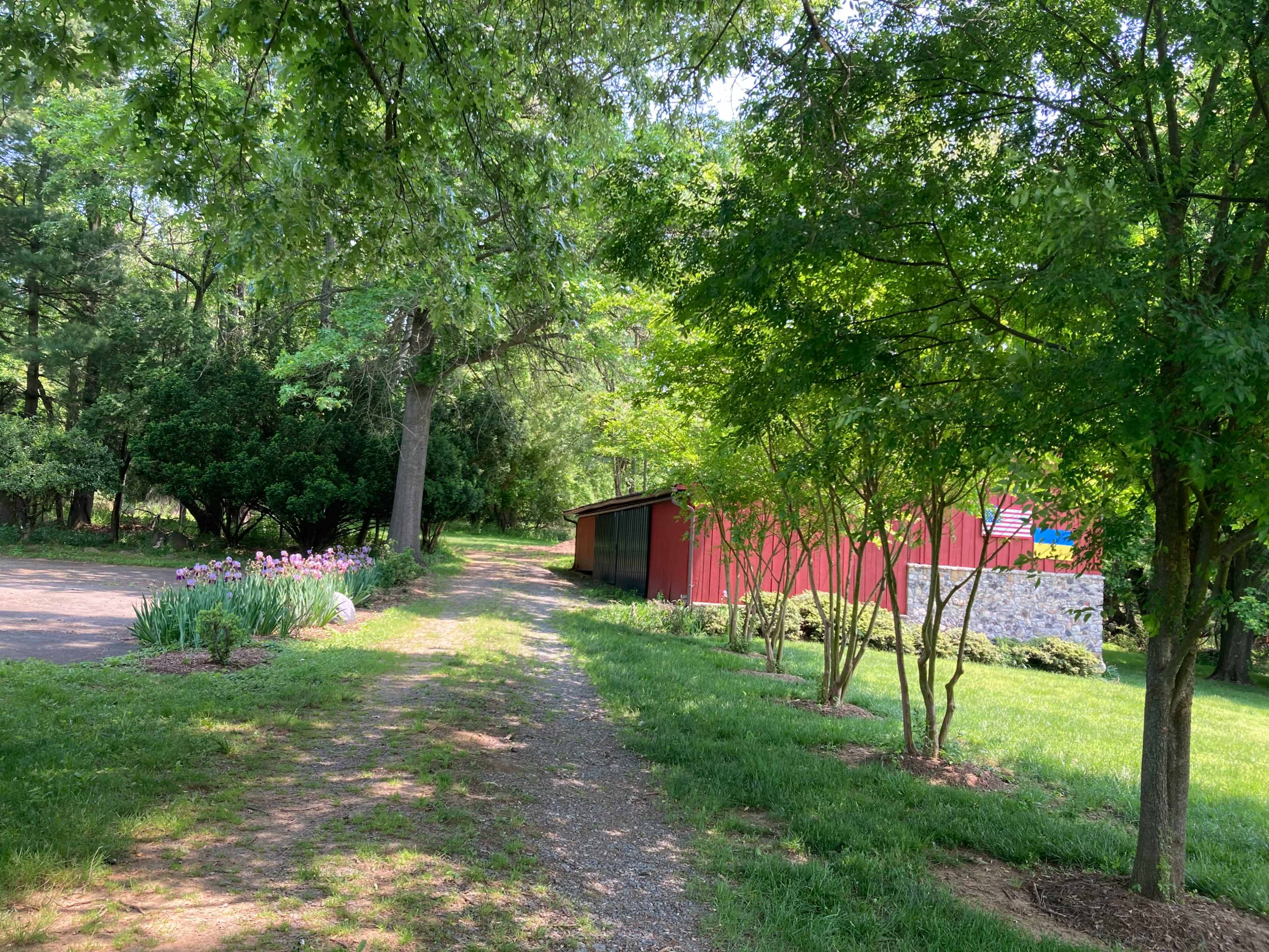 A gravel path leads between lush greenery to a red barn surrounded by trees and flower beds.