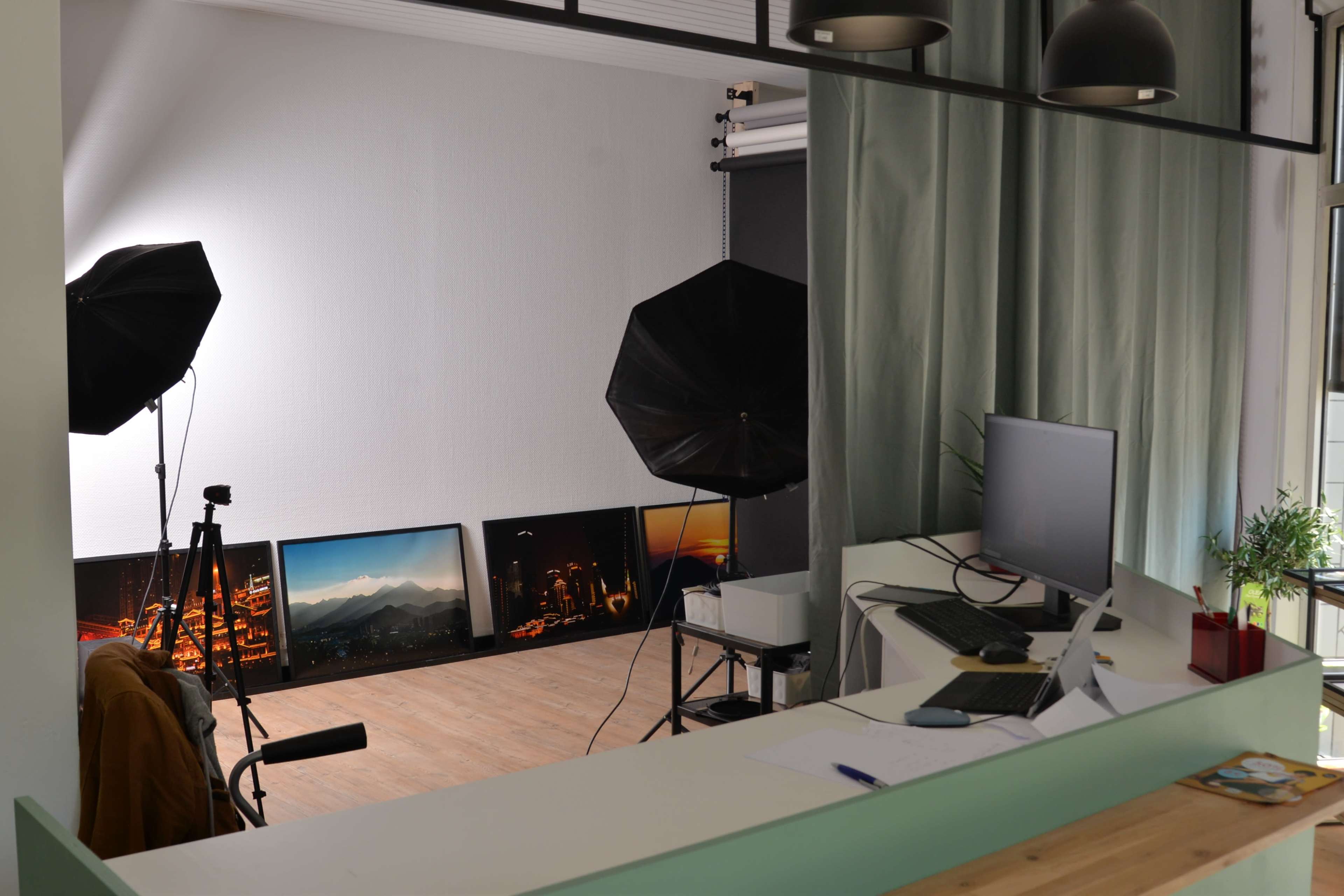 A photography studio with softbox lights, a camera on a tripod, artwork displayed on the floor, and a workstation with a computer on a counter.