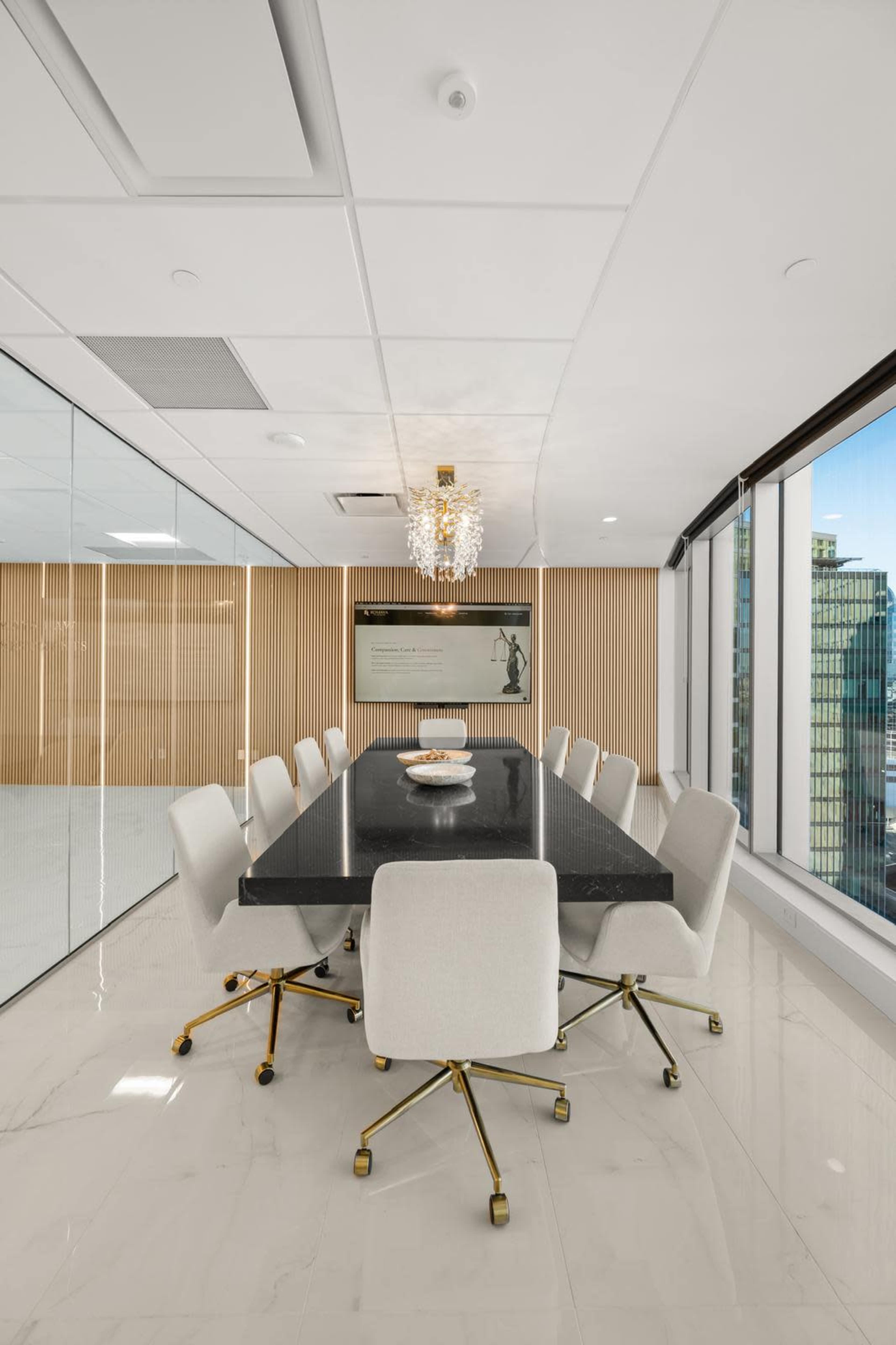 A modern conference room features a large black table surrounded by white chairs, with a chandelier hanging above and large windows offering a view of the city.