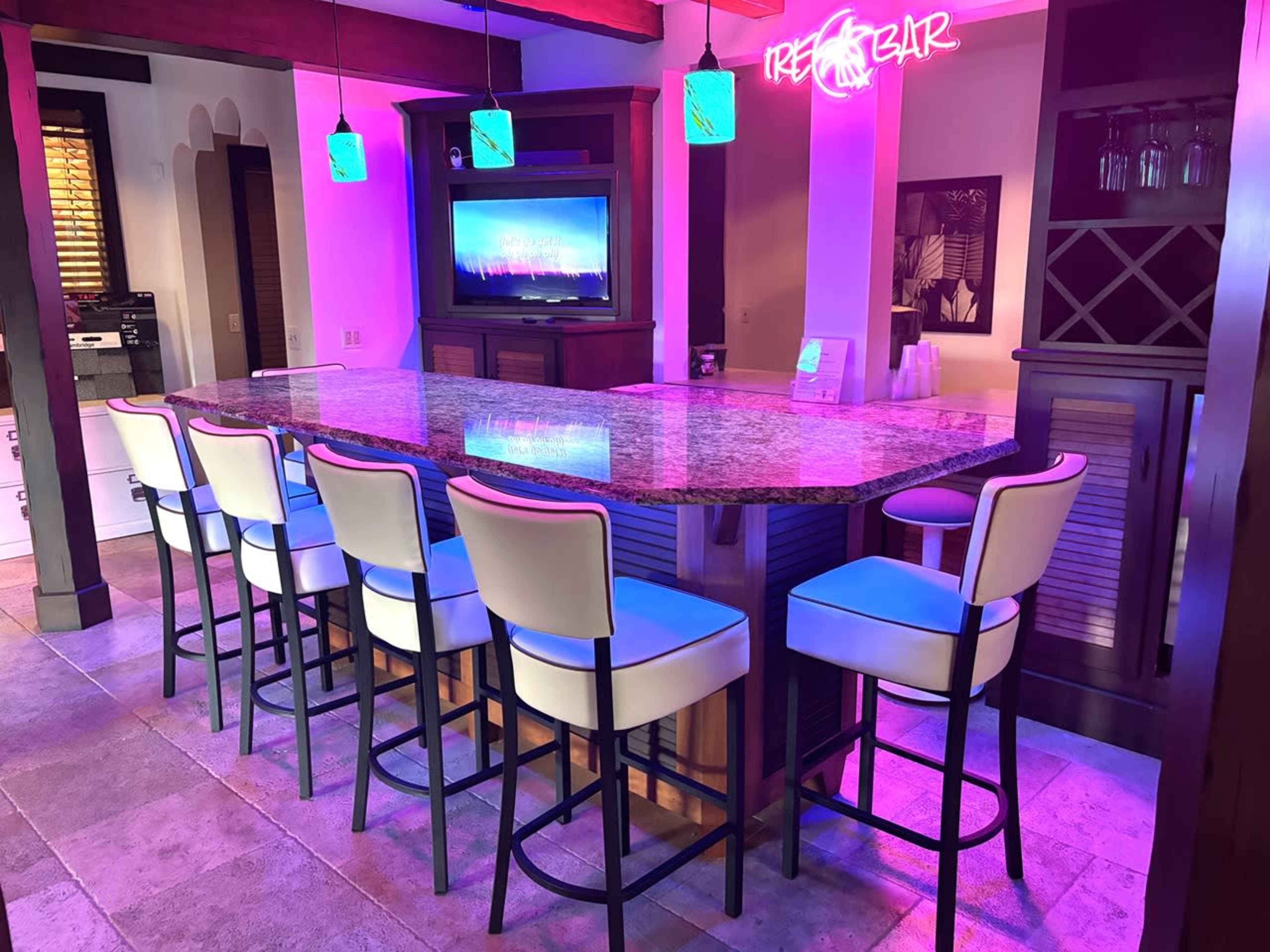 A modern home bar features a curved granite countertop with eight white bar stools and a neon sign, illuminated by colorful lights.