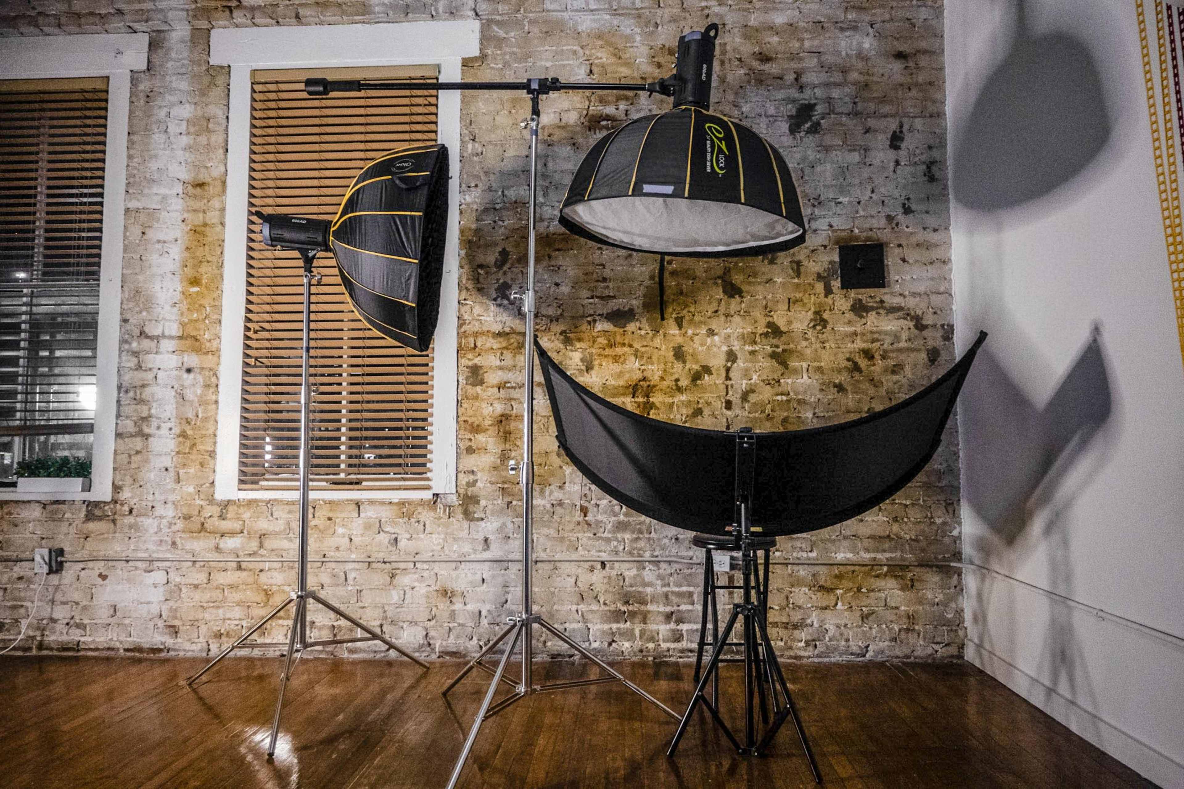 Three lighting setups in a studio: two softboxes on stands and a reflector positioned on a tripod, against a backdrop of exposed brick walls and wooden flooring.