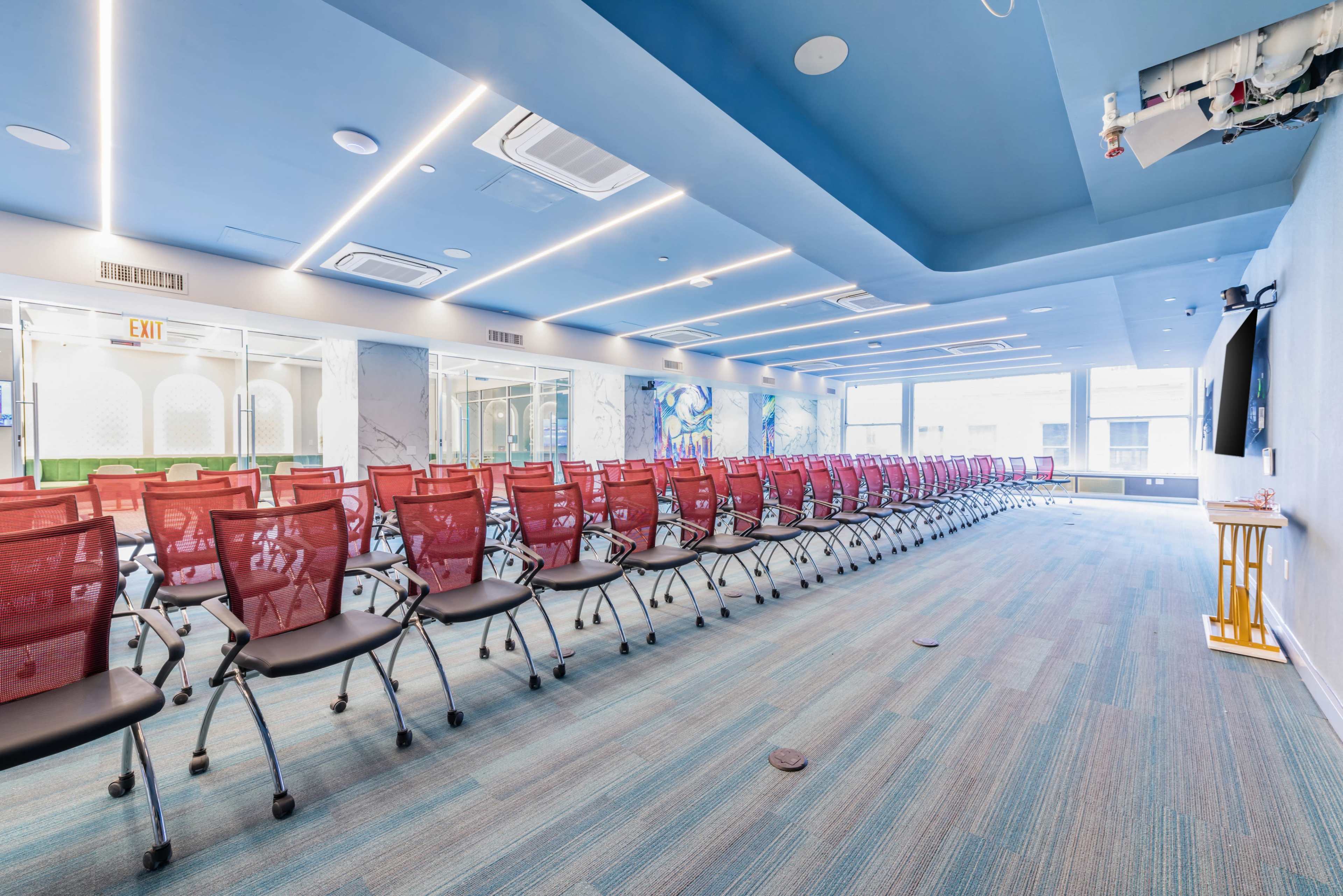 The image shows a spacious conference room arranged with rows of red chairs facing a large screen, illuminated by ceiling lights and featuring a blue ceiling.