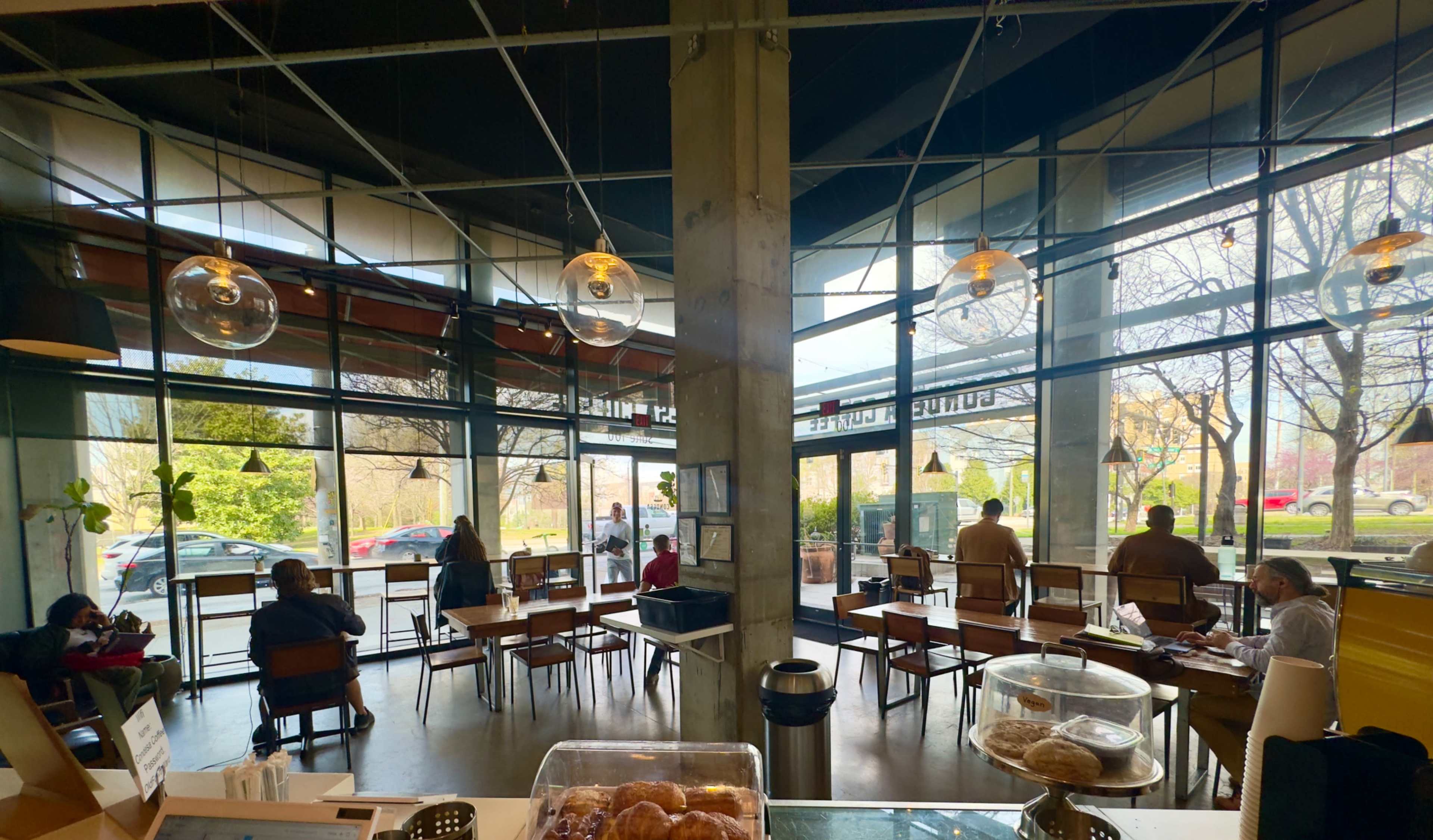 The image shows a café interior with large windows, several tables occupied by patrons, and a countertop displaying baked goods.
