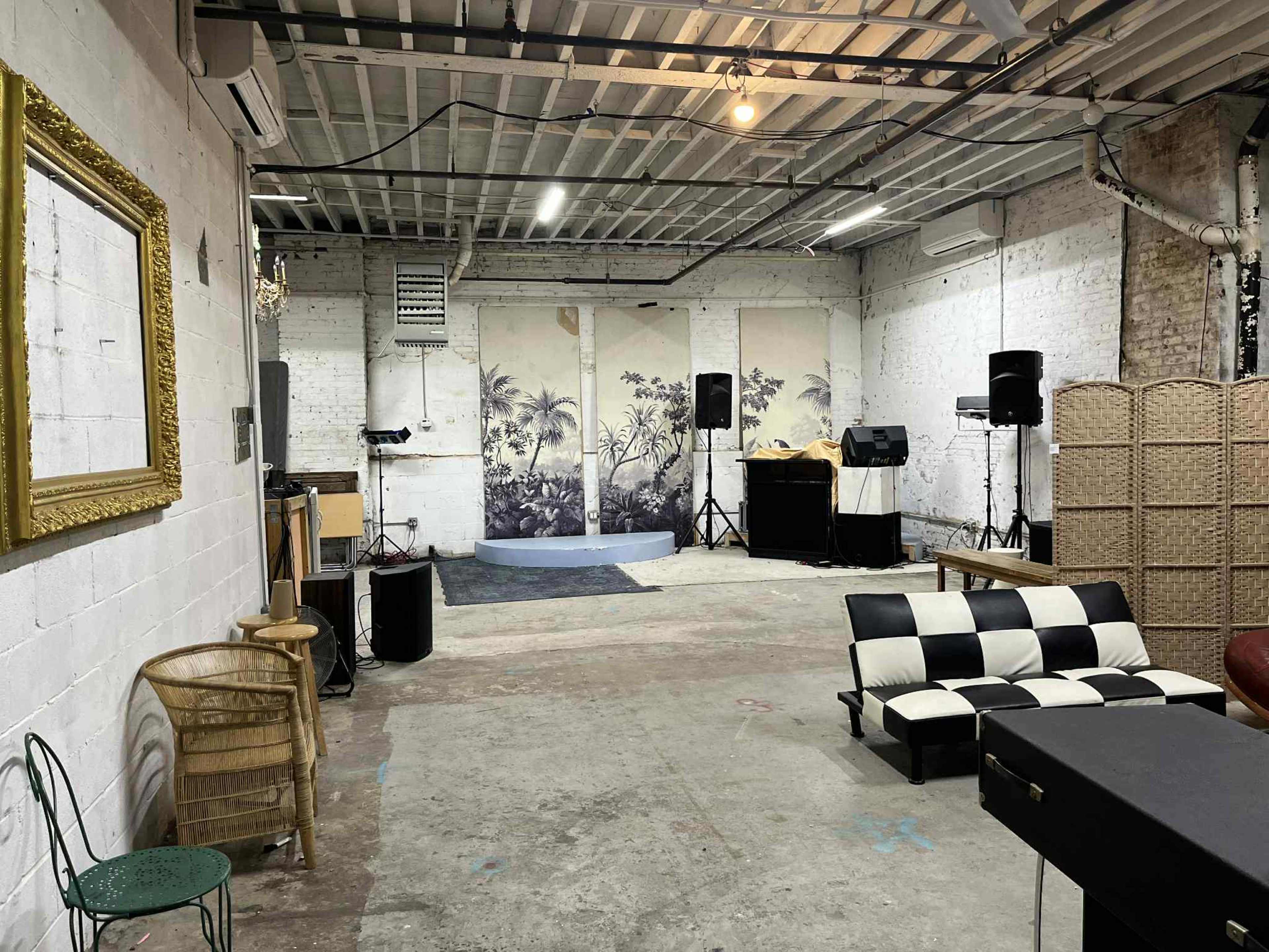 The image shows an empty industrial-style room with a stage, speakers, and a mix of seating including a checkered couch and a woven screen in the background.