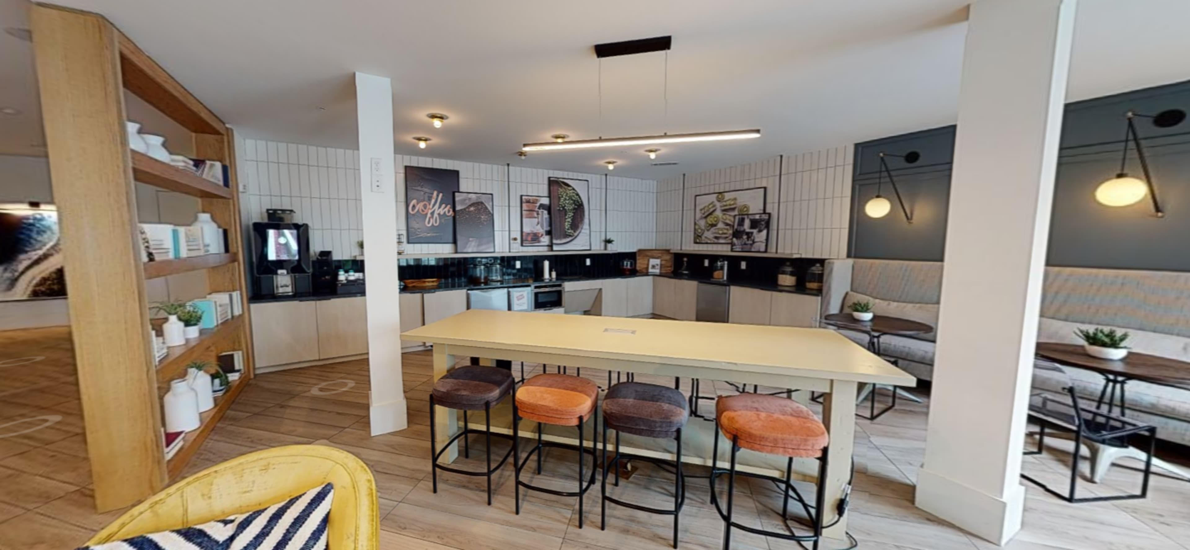 The image shows a modern kitchen area with a central yellow table, bar stools, and a wall-mounted shelf containing various items.