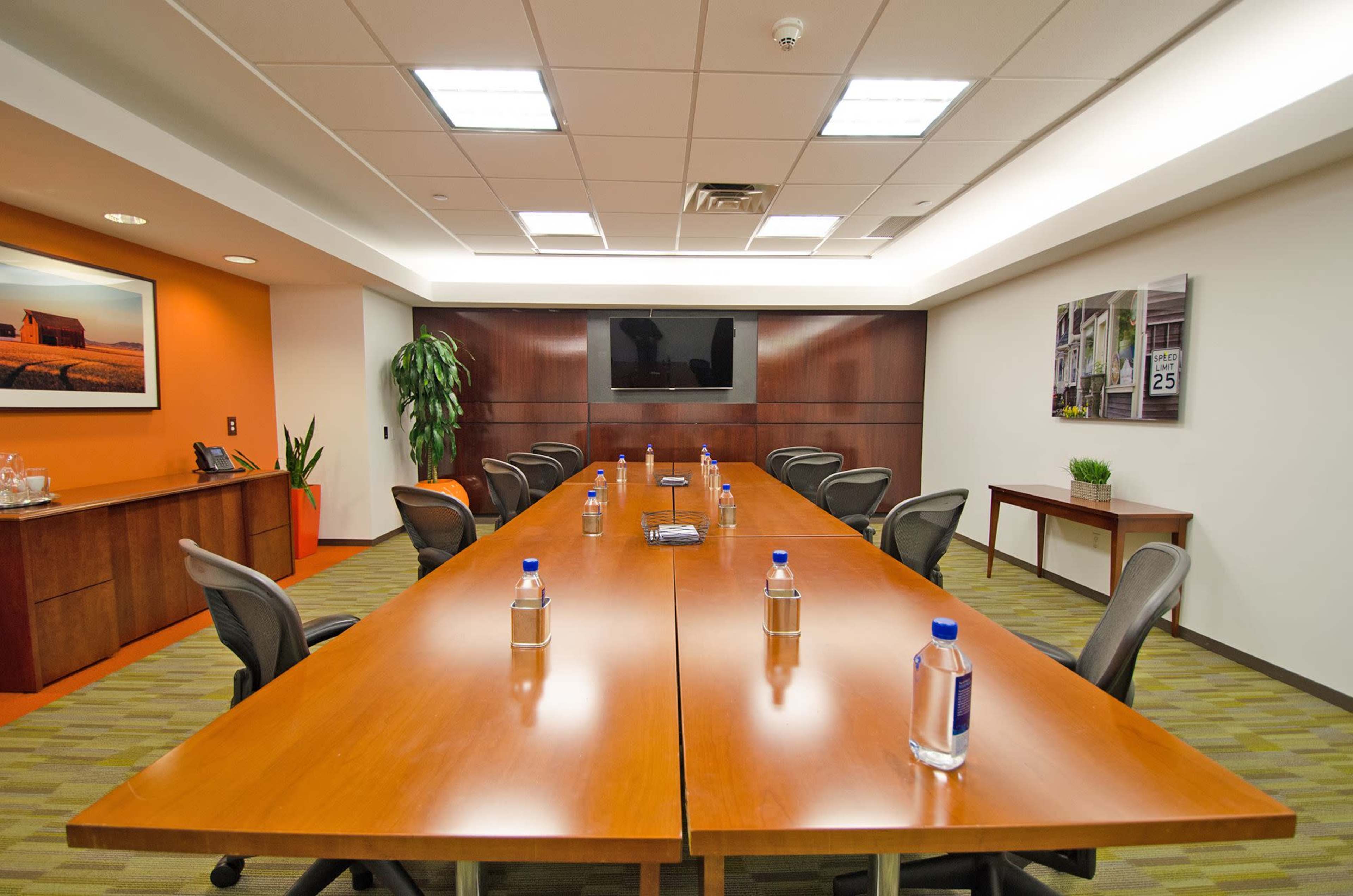 A long wooden conference table is set with water bottles and surrounded by office chairs in a well-lit meeting room with a television and decorative plants.