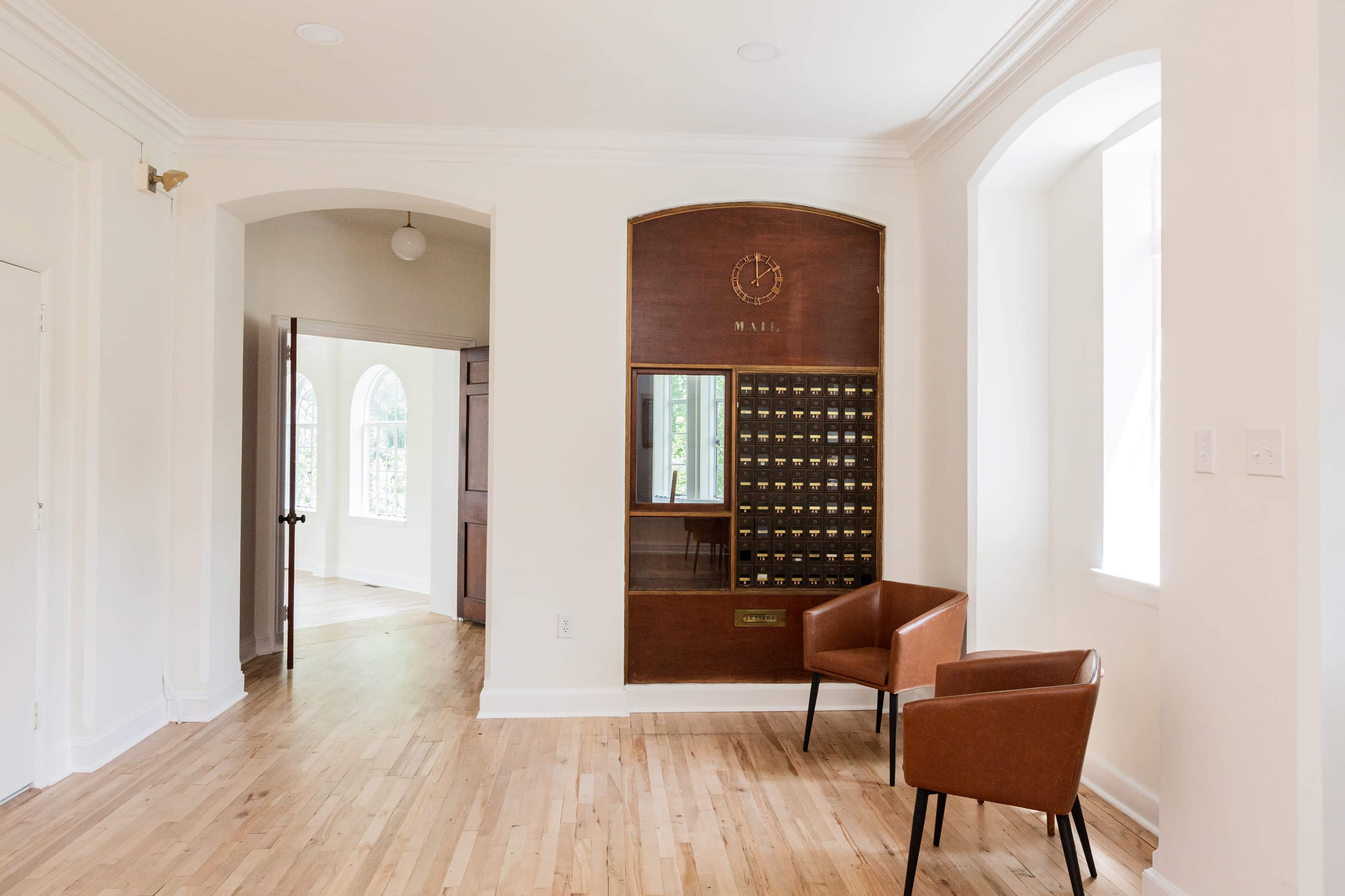 A minimalist entryway featuring a wooden mail slot with a clock above it, two brown chairs, and an open doorway leading to another room.
