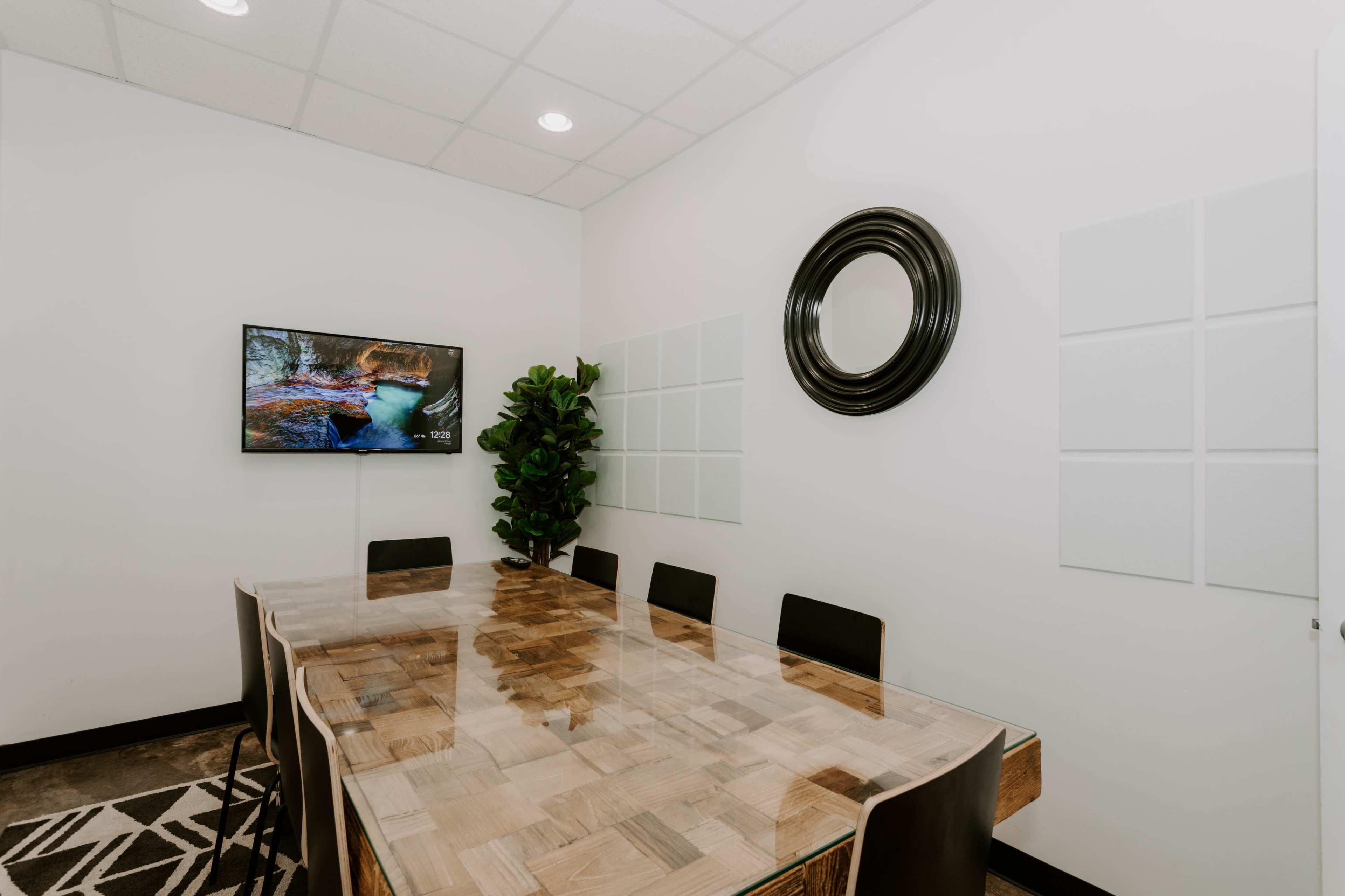 A meeting room features a wooden table surrounded by black chairs, a wall-mounted TV, and a decorative plant.