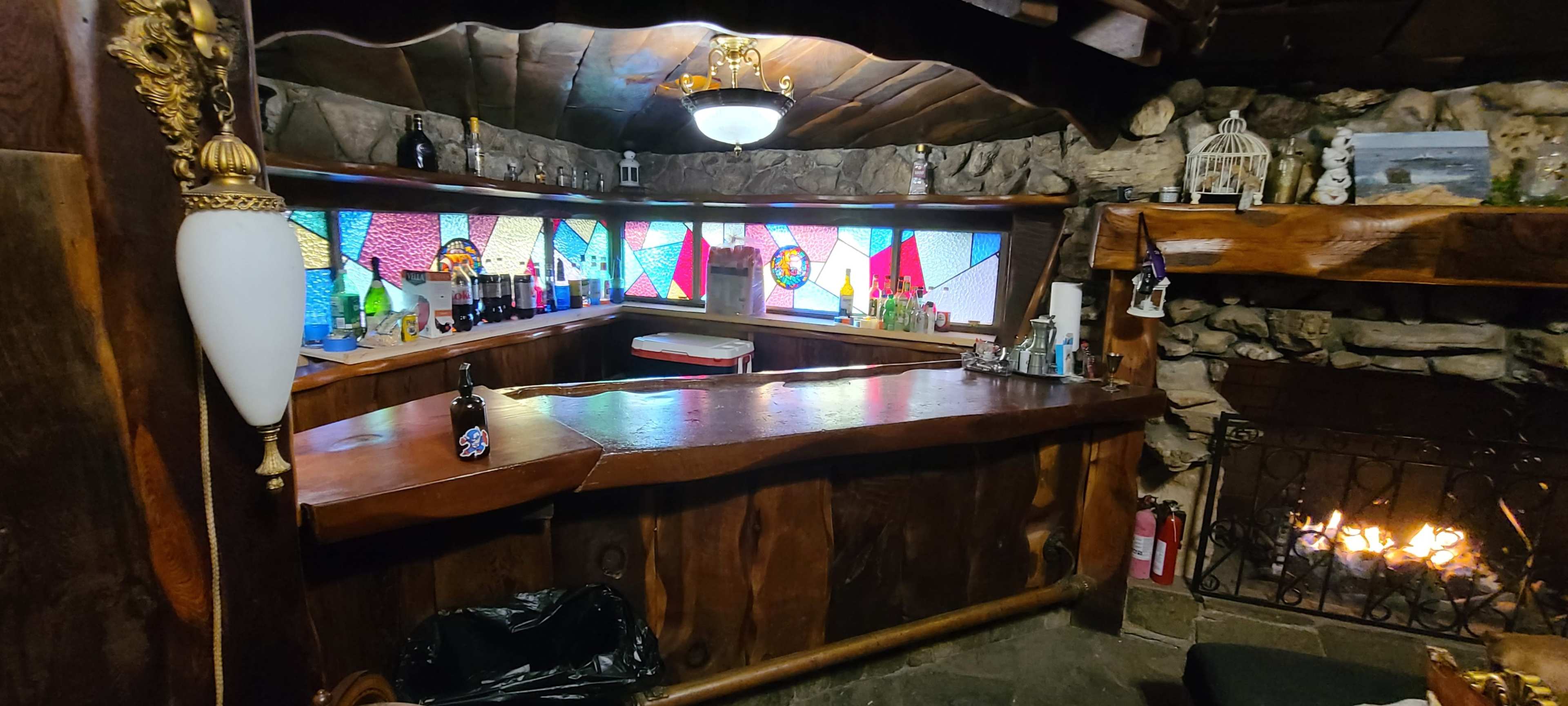 A rustic bar area with a wooden counter, bottles displayed on the shelves, and a stone fireplace with a fire burning.
