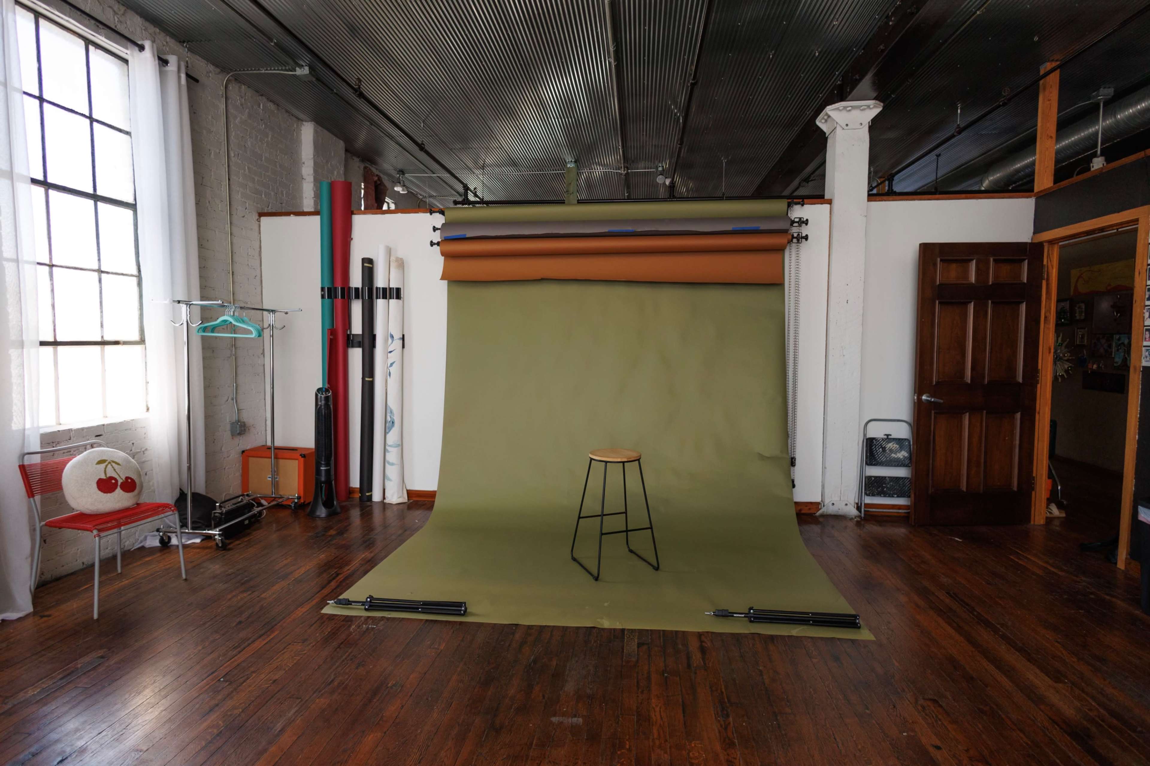 The image shows a photography studio with a green backdrop, a stool in the center, and various equipment and props arranged around the space.