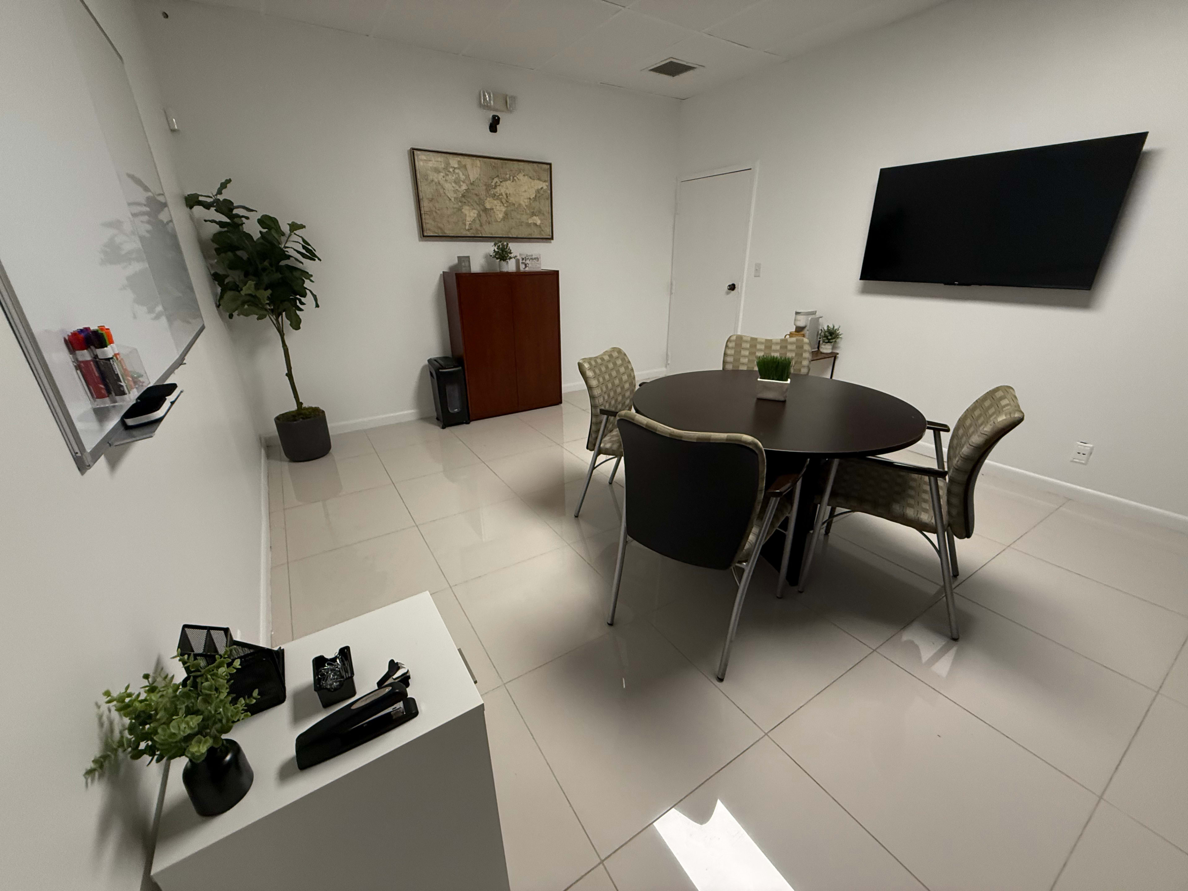 The image shows a modern meeting room with a round table, four chairs, a whiteboard, a wall-mounted TV, and a map on the wall.