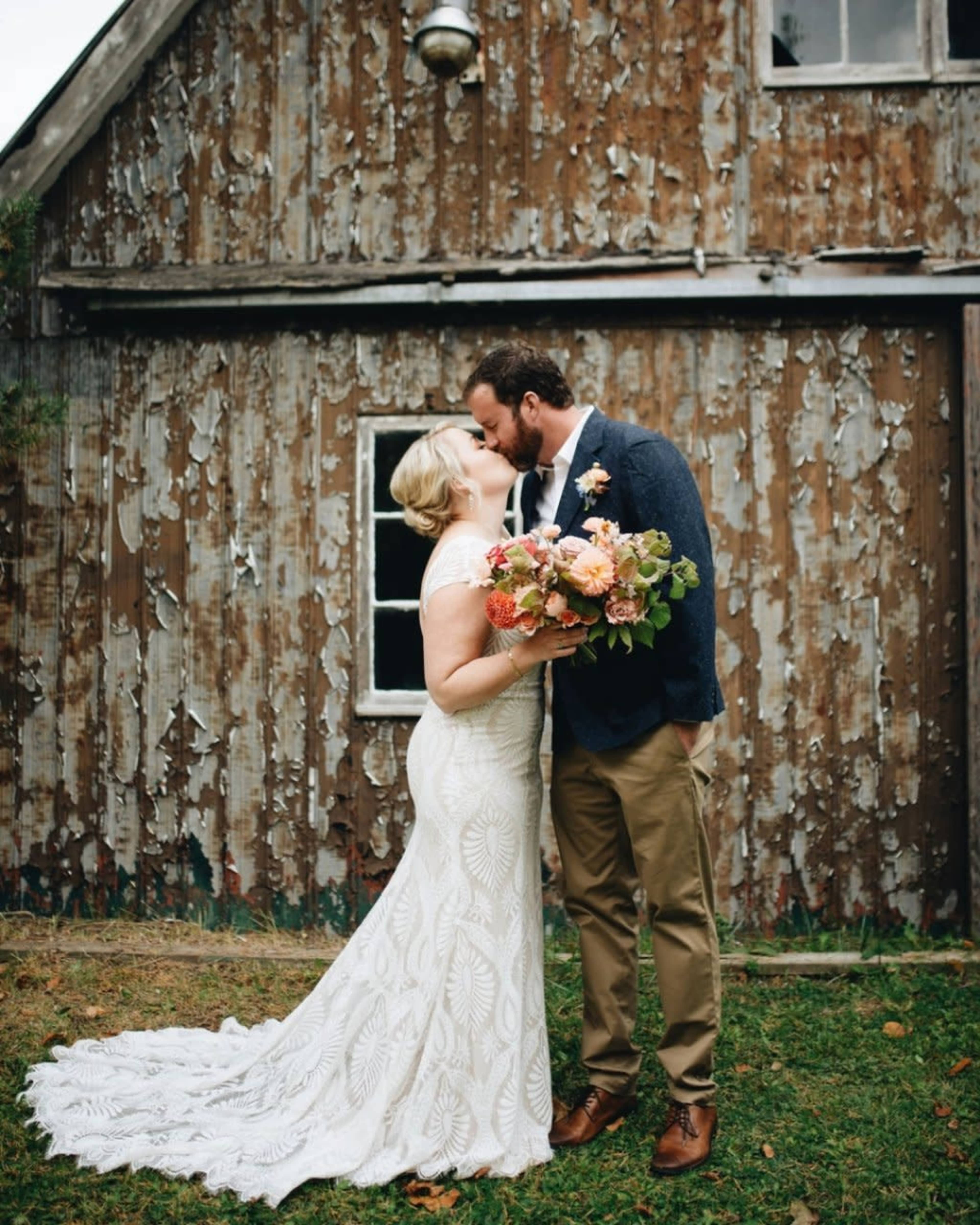 A bride in a lace wedding dress kisses a groom in a suit outside a rustic, weathered building.