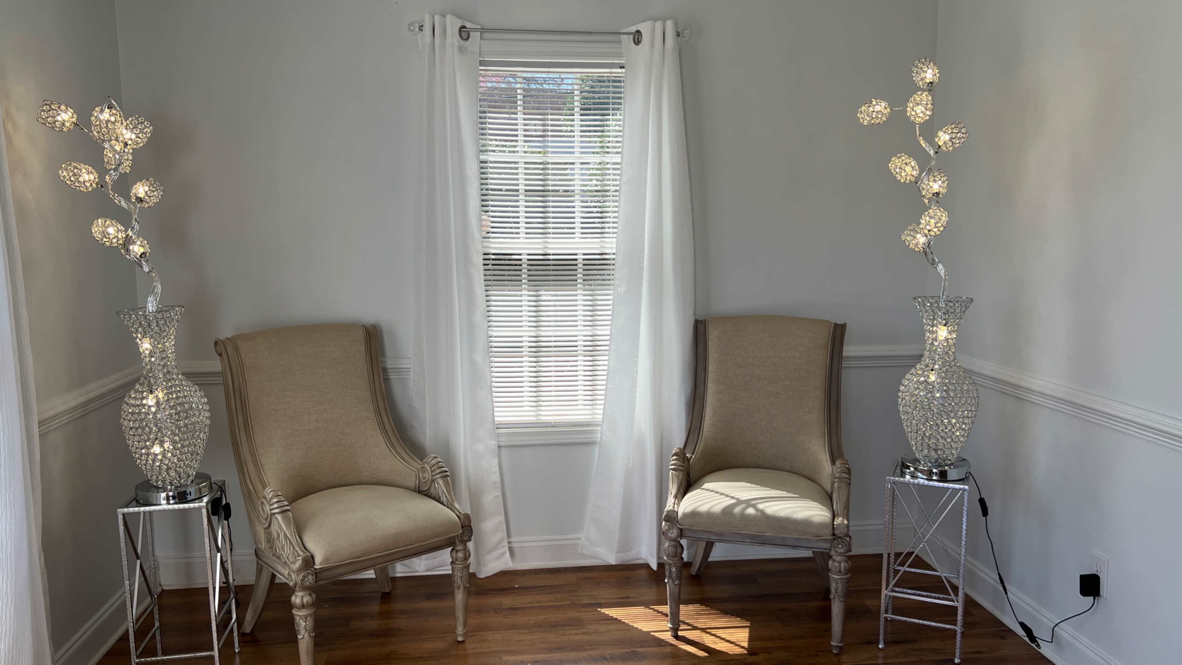 Two upholstered chairs are positioned on either side of a window, with decorative vases containing lights next to them.