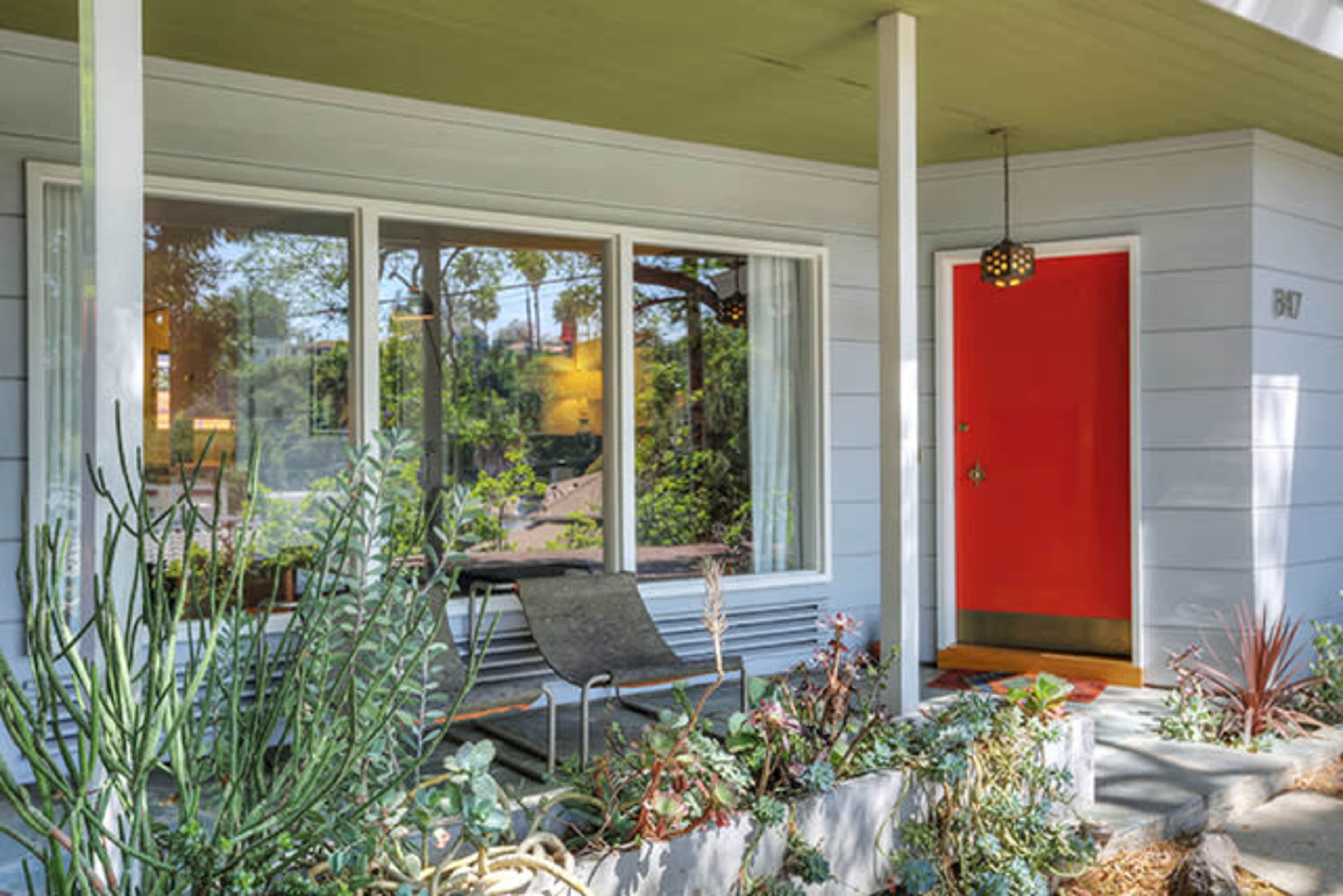 A modern house facade with a bright red door, large front windows, and a small seating area surrounded by plants.