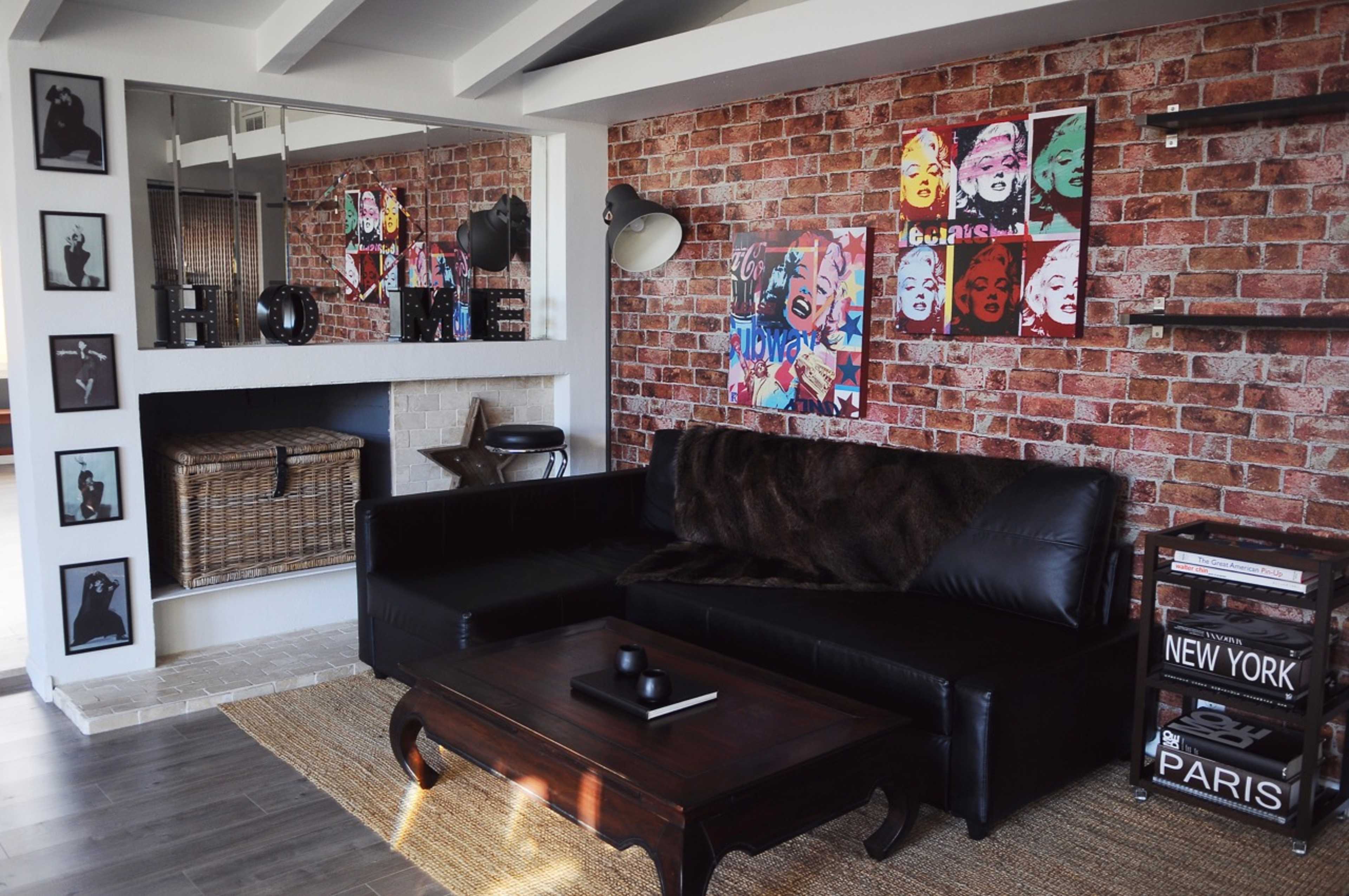 The image shows a modern living room featuring a black leather sofa, a wooden coffee table, and a wall adorned with colorful pop art alongside exposed brick.