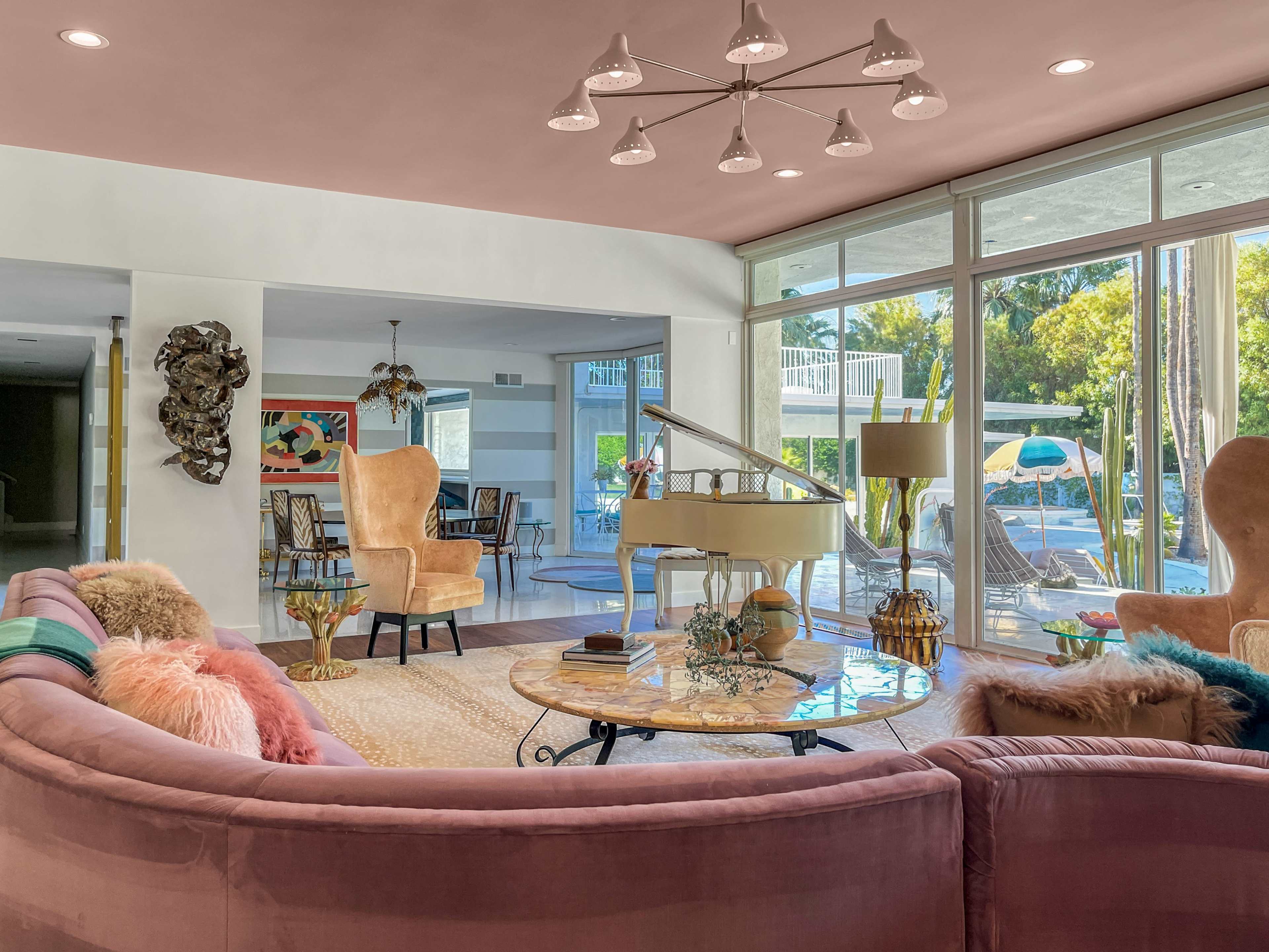 A modern living room with a large pink sofa, a glass coffee table, a grand piano, and large windows overlooking a pool area.