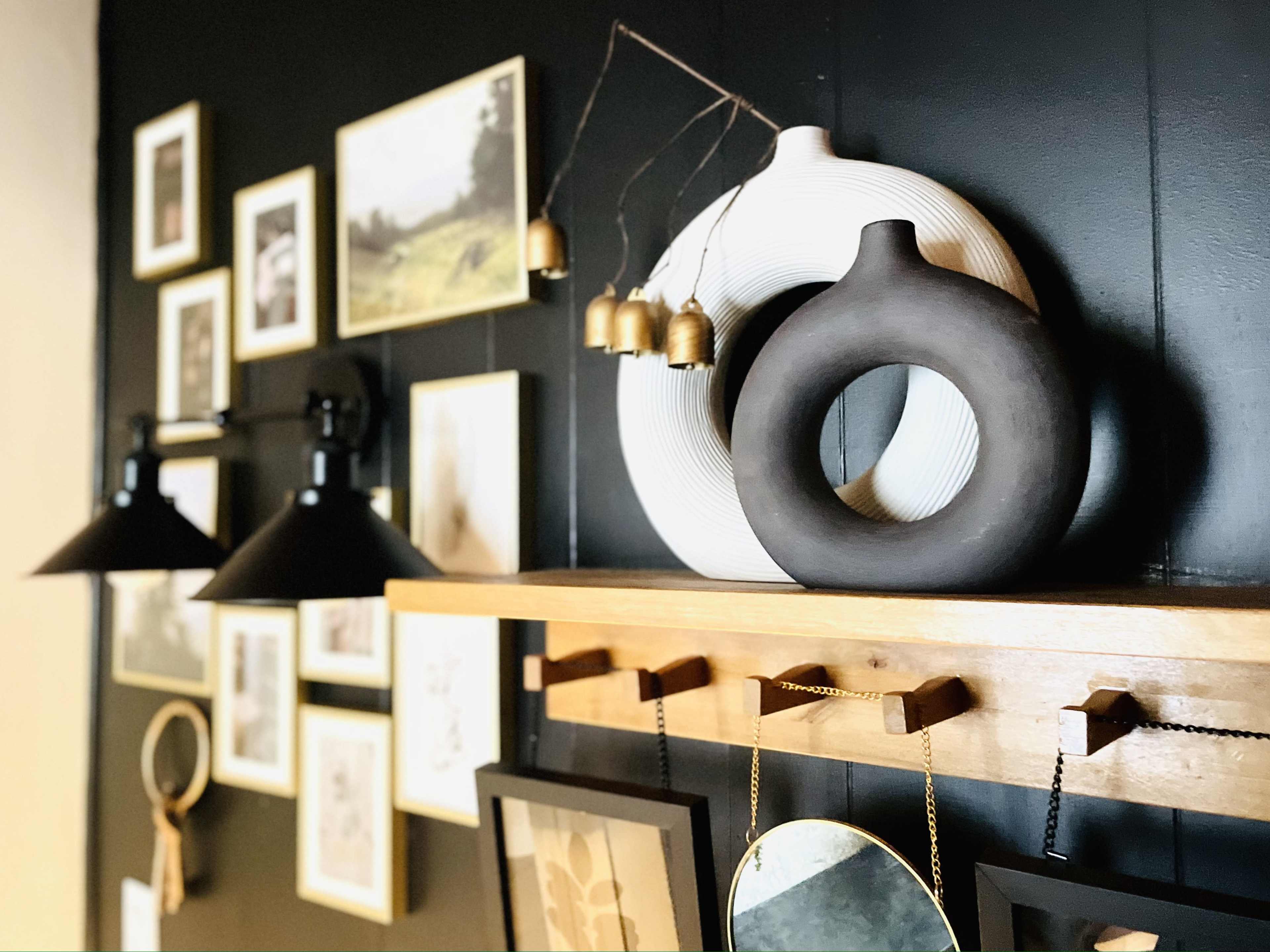 The image features a mounted shelf with decorative objects, including a white circular vase and a dark round vase, against a wall adorned with framed photographs and artwork.